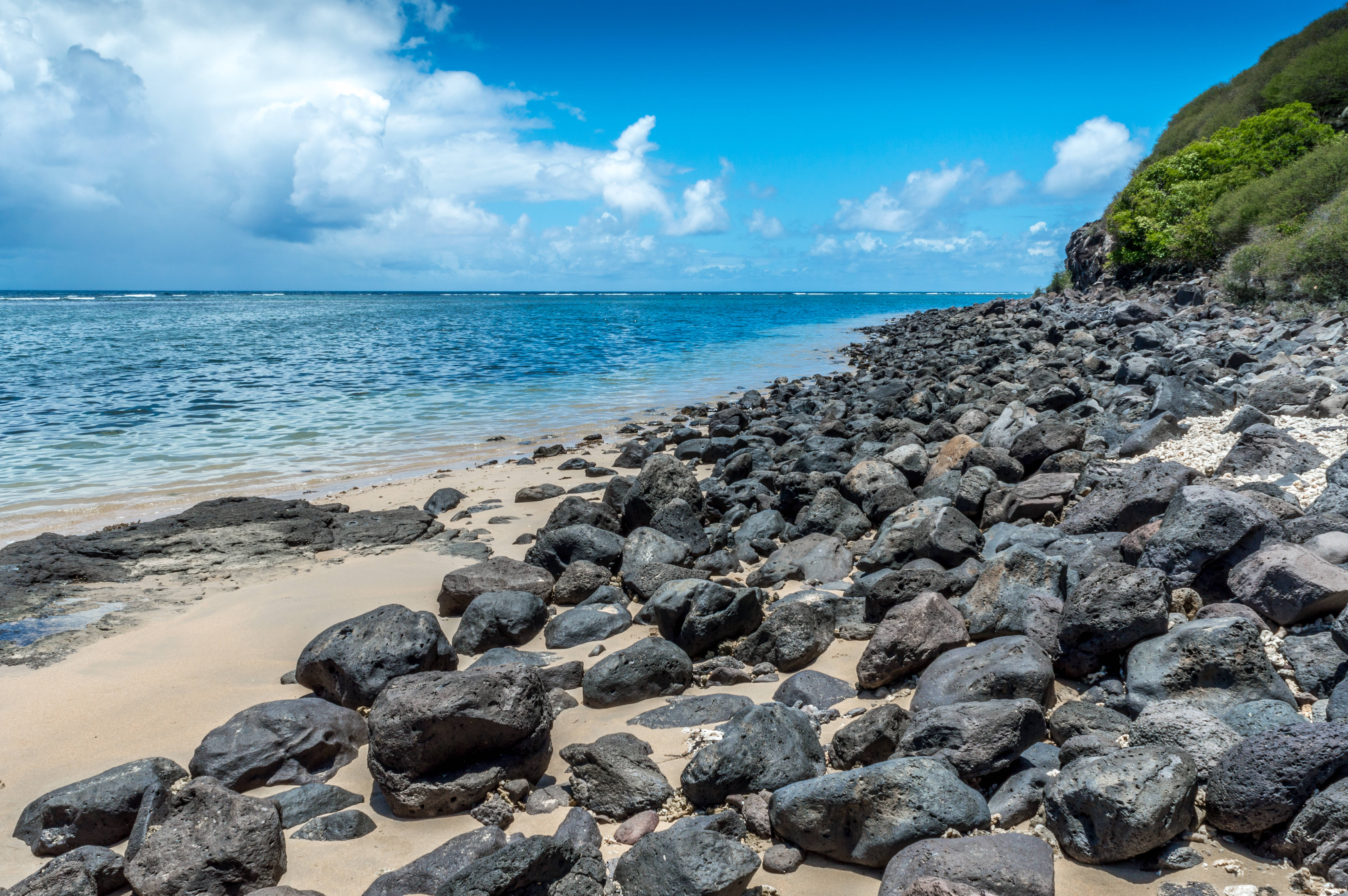 Mauritius 桌布 壁紙 — 一個陽光明媚的岩石海灘，深色火山石散落在金色沙灘上，與碧綠的海洋相接，上方是明亮的藍天和蓬鬆的白雲。