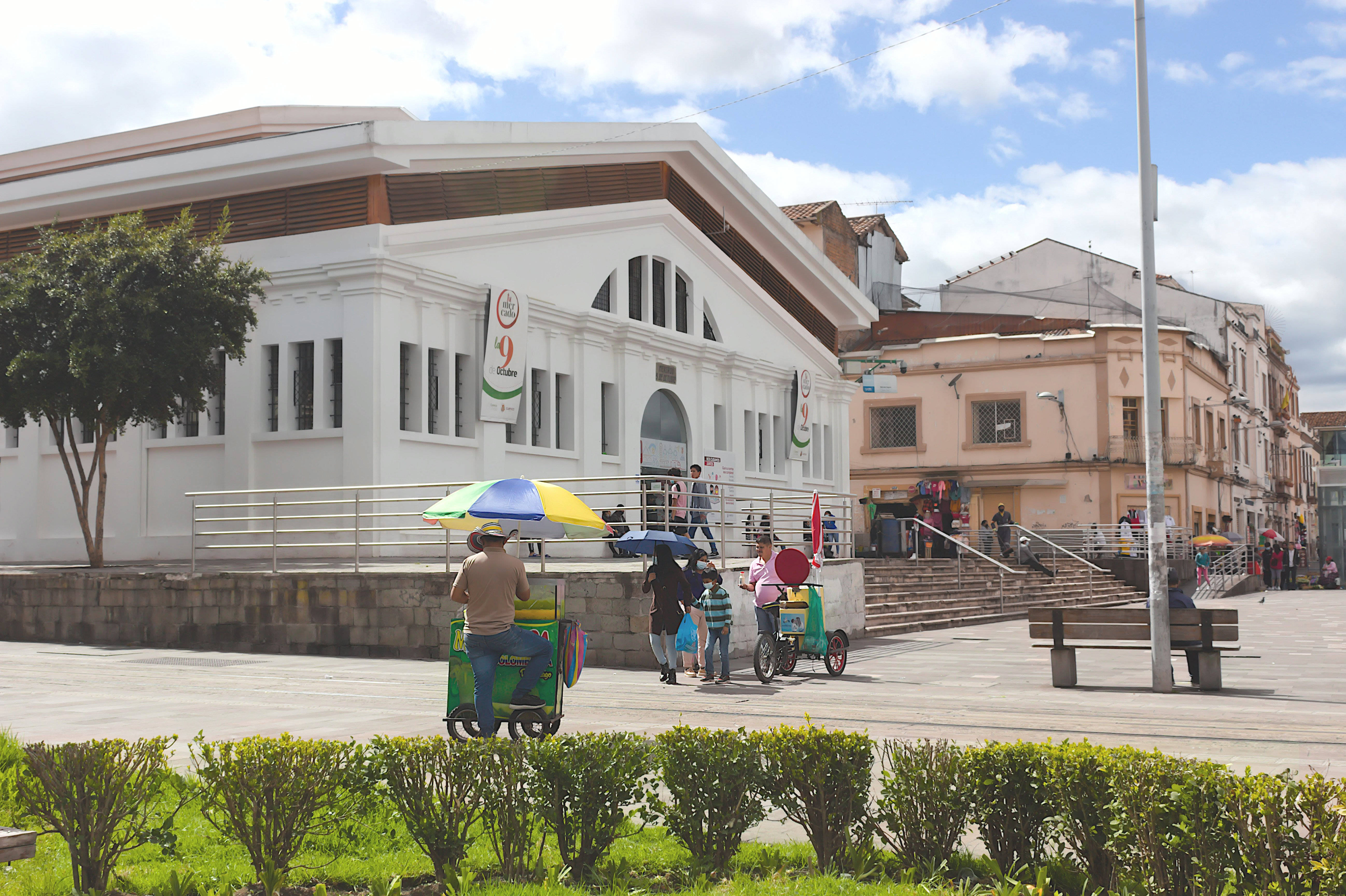 Ecuador 桌布 壁紙 — 一個陽光明媚的廣場場景，有色彩繽紛的手推車小販和行人，靠近一棟帶有拱形窗戶的白色建築。
