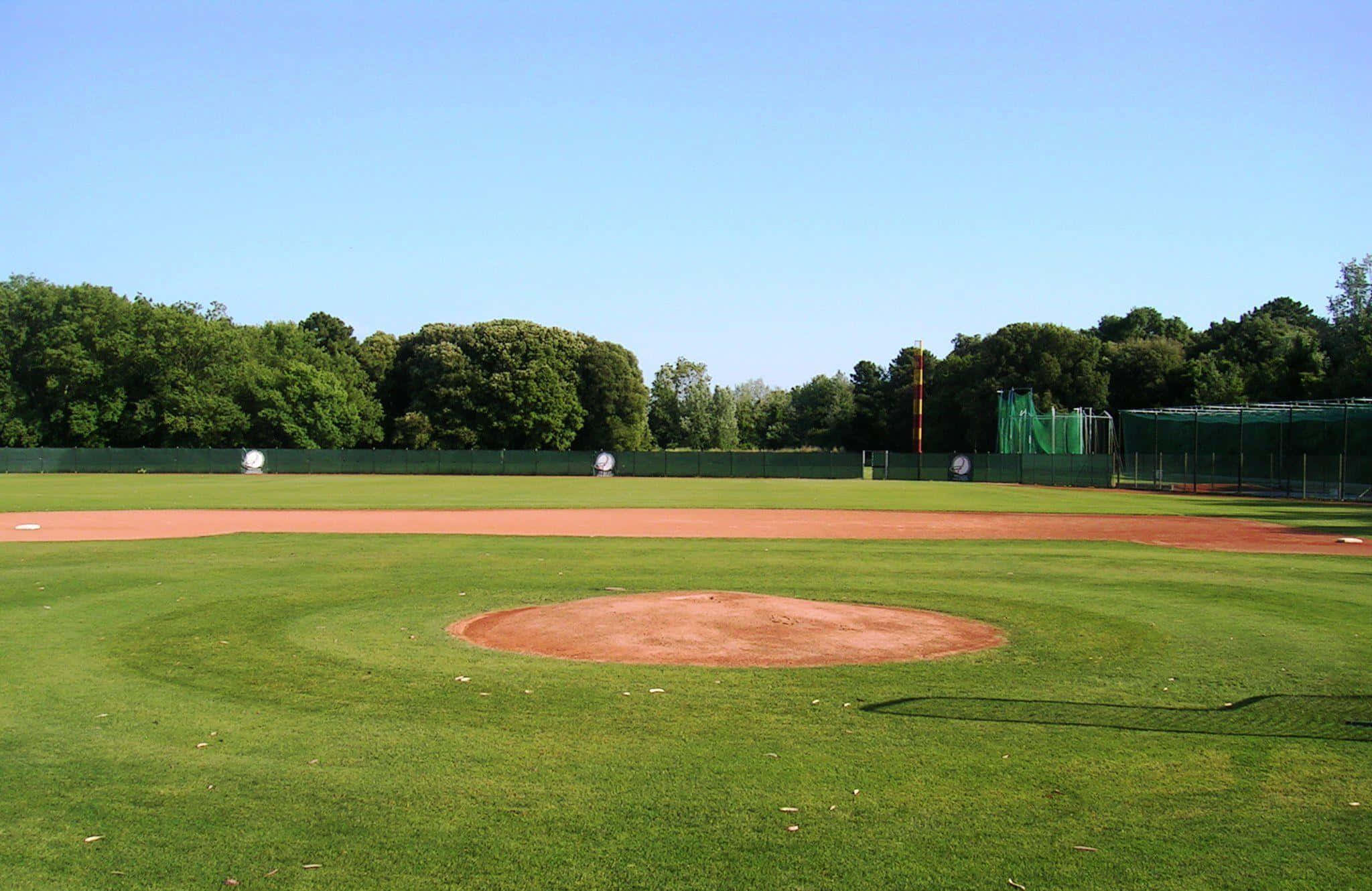 Baseball Field 桌布 壁紙 — 一個陽光明媚的棒球場，在清澈的藍天下，有充滿活力的綠色草地外野和紅褐色的投手丘。