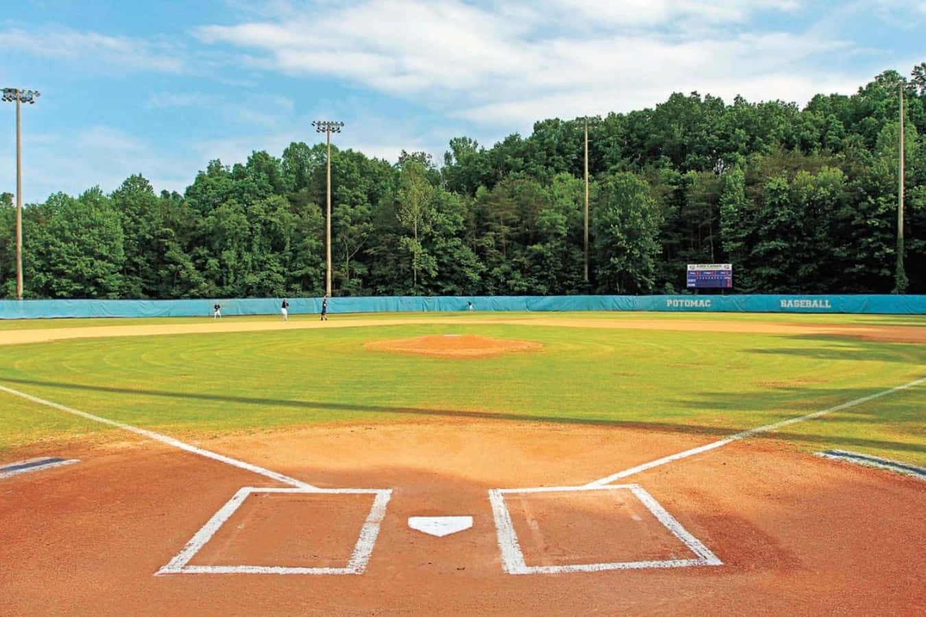 Baseball Field 桌布 壁紙 — 一個陽光明媚的棒球場，有綠色草地和棕色泥土，被茂密的森林環繞，上方是明亮的藍天和散落的雲朵。