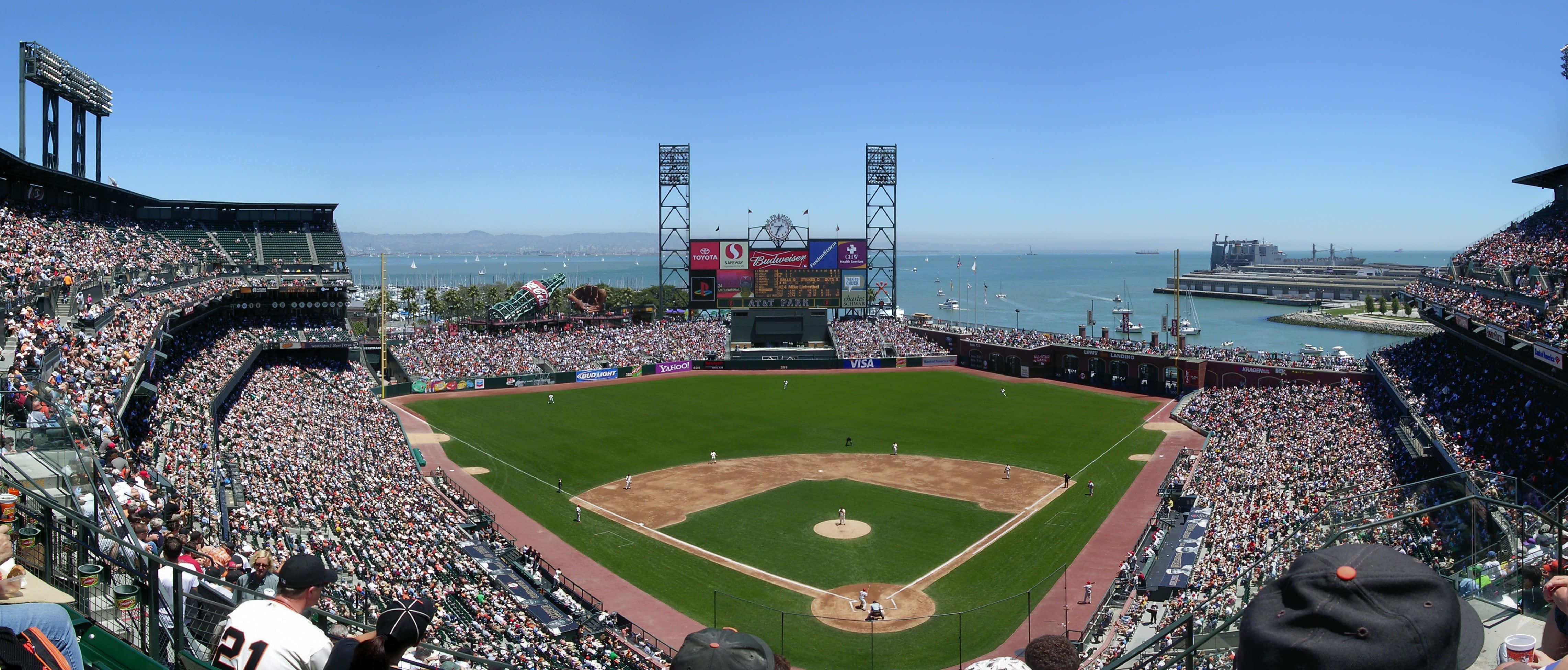 Baseball Stadium 桌布 壁紙 — 一個陽光明媚的棒球場全景，擠滿了球迷，俯瞰著一個有帆船的海灣。