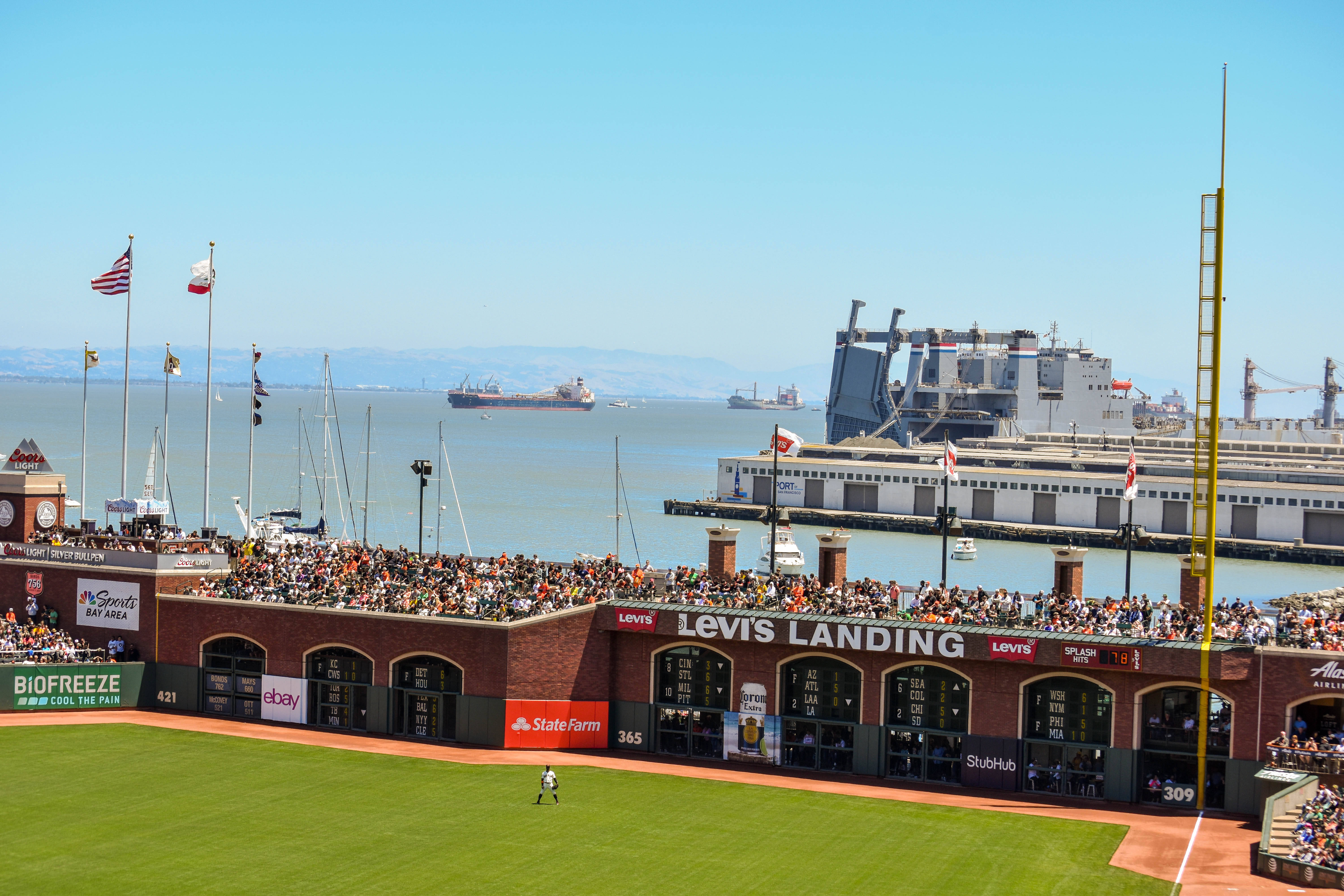 San Francisco Giants 桌布 壁紙 — 一個陽光明媚的棒球場擠滿了球迷，俯瞰著一個有船隻和大型造船廠的港口，天空湛藍。
