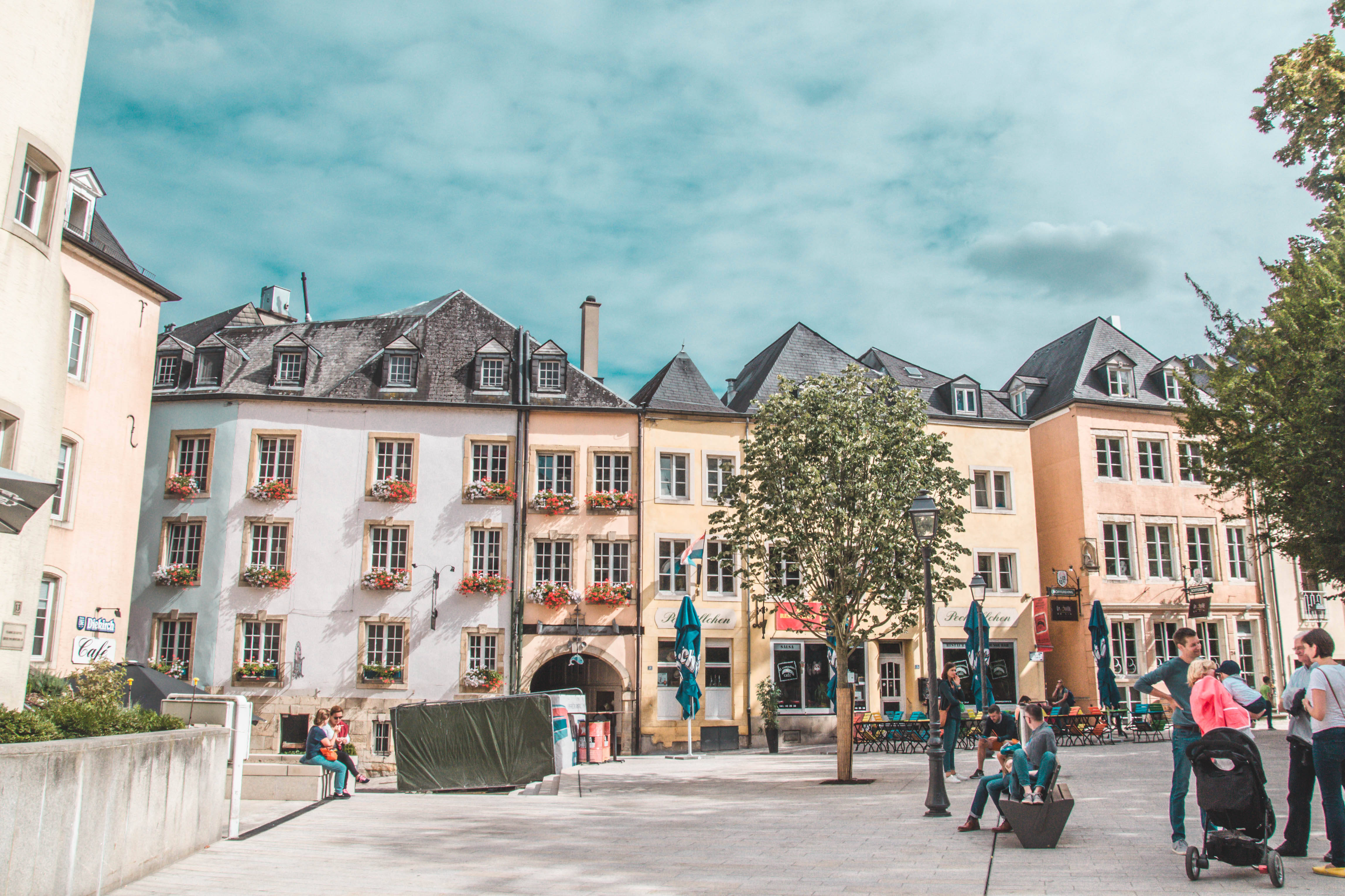 Luxembourg 桌布 壁紙 — 一個陽光明媚的歐洲城鎮廣場，有柔和色調的建築、花箱，人們在部分多雲的天空下在戶外咖啡館放鬆。