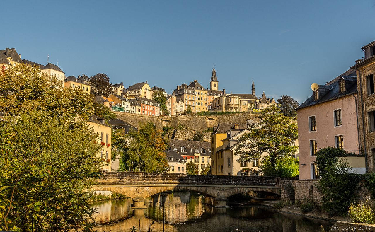 Luxembourg 桌布 壁紙 — 一個陽光明媚的歐洲小鎮，歷史建築坐落在寧靜河流上方的山坡上，並有一座石橋。