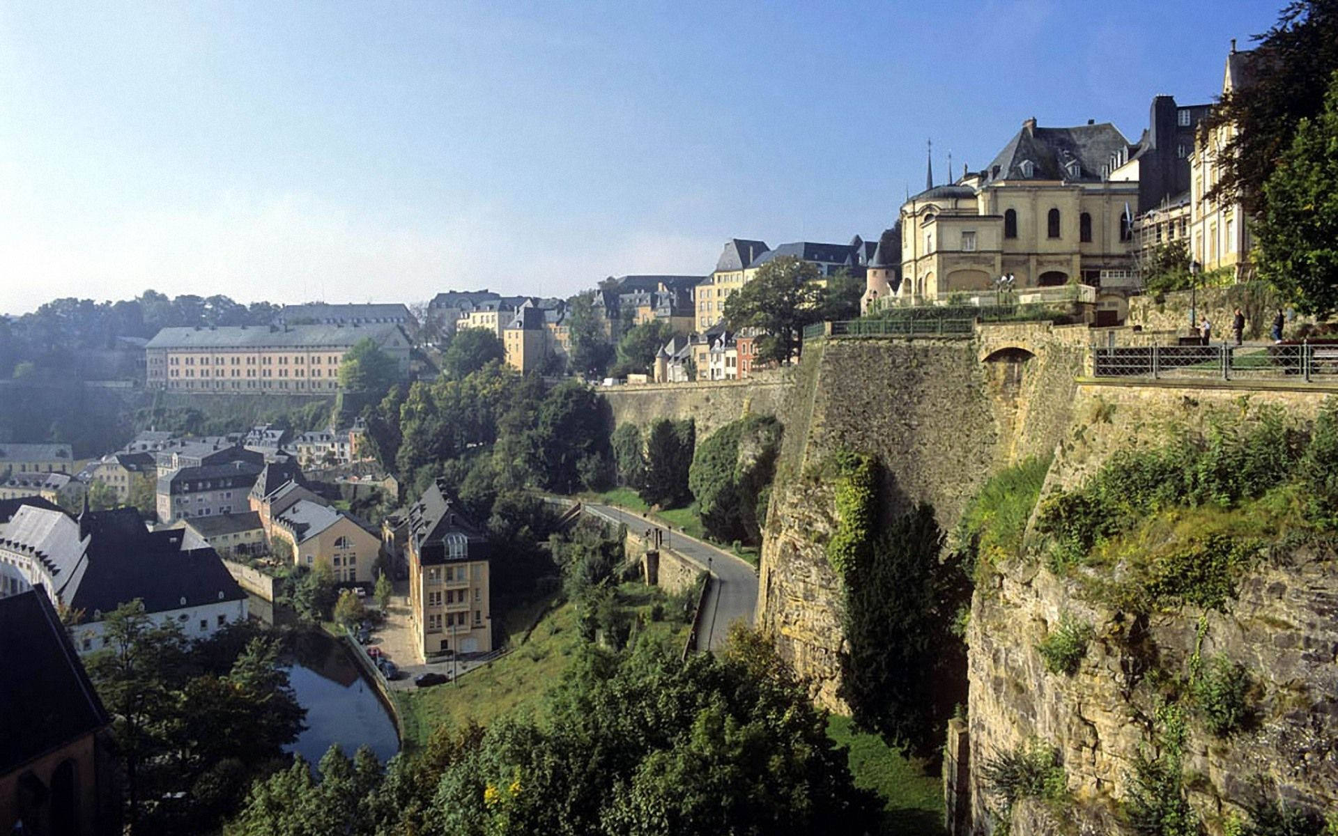 Luxembourg 桌布 壁紙 — 一個陽光明媚的歐洲小鎮依附在陡峭的懸崖邊，擁有歷史悠久的石頭建築和蜿蜒的道路，坐落在茂密的綠樹之中。