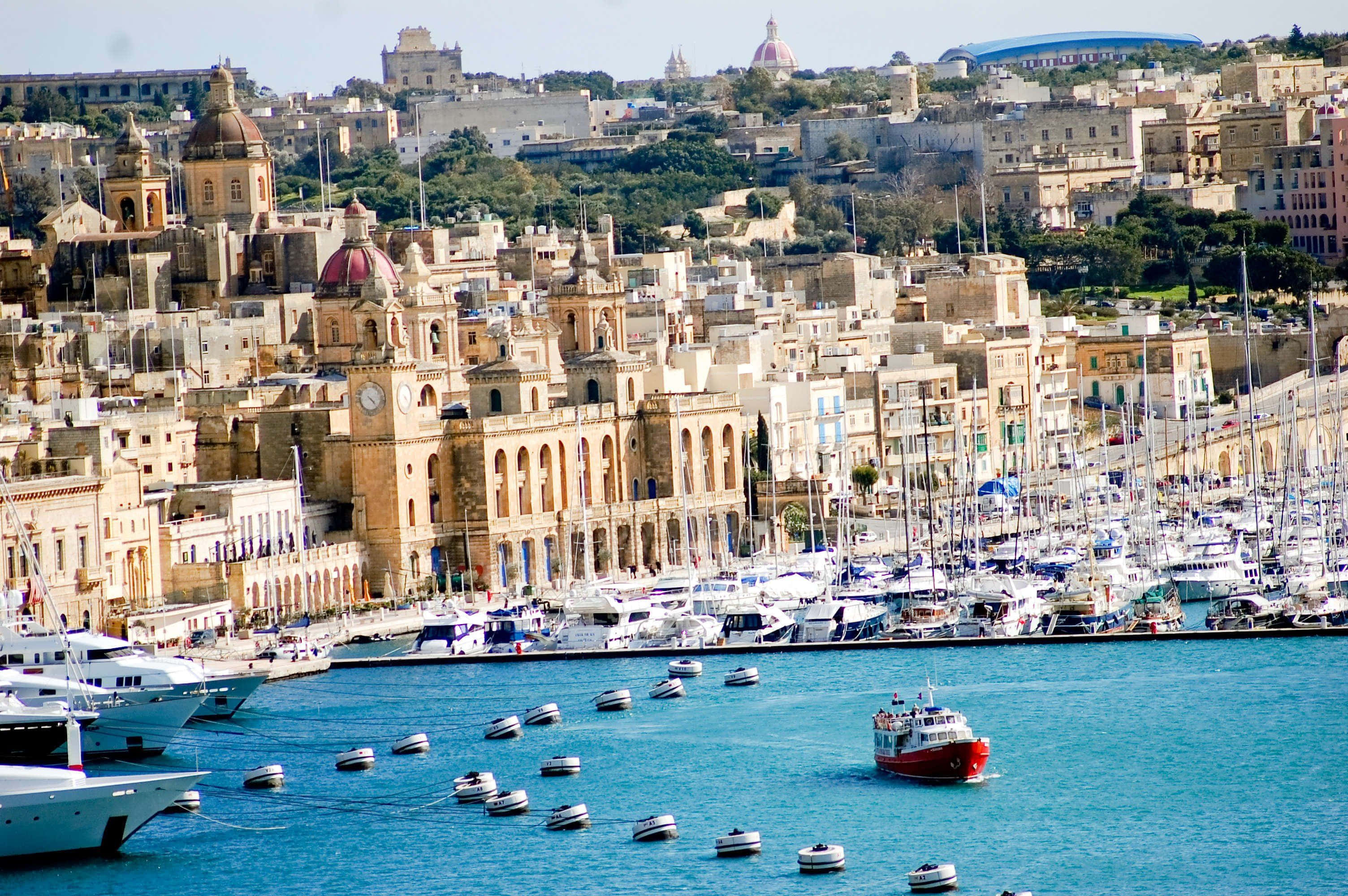 Malta 桌布 壁紙 — 一個陽光明媚的港口，停滿了船隻和遊艇，依偎在擁有圓頂教堂的歷史石建築山坡旁。