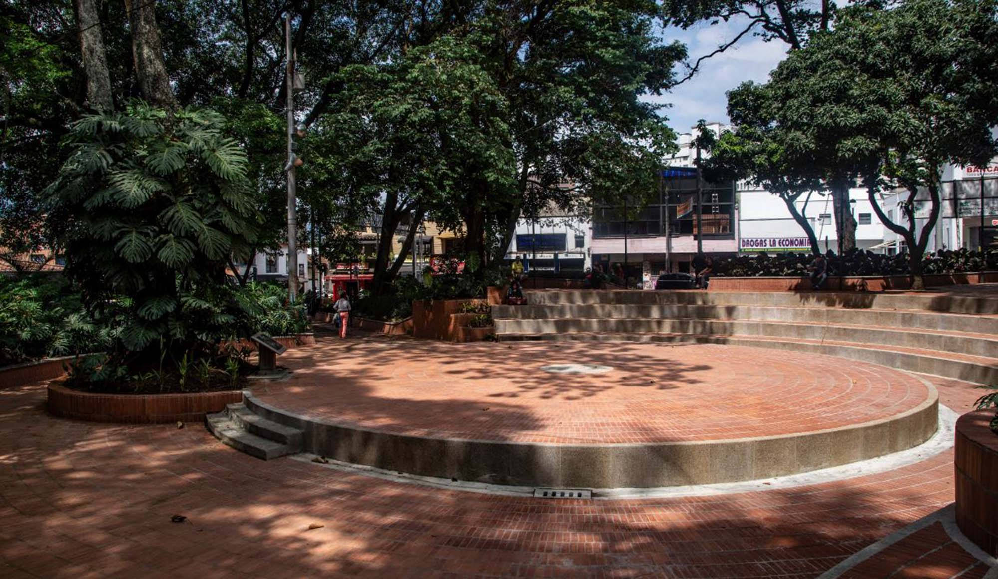 Medellin 桌布 壁紙 — 一個陽光普照的圓形磚砌廣場，周圍環繞著茂密的綠樹和城市建築，投下斑駁的陰影。