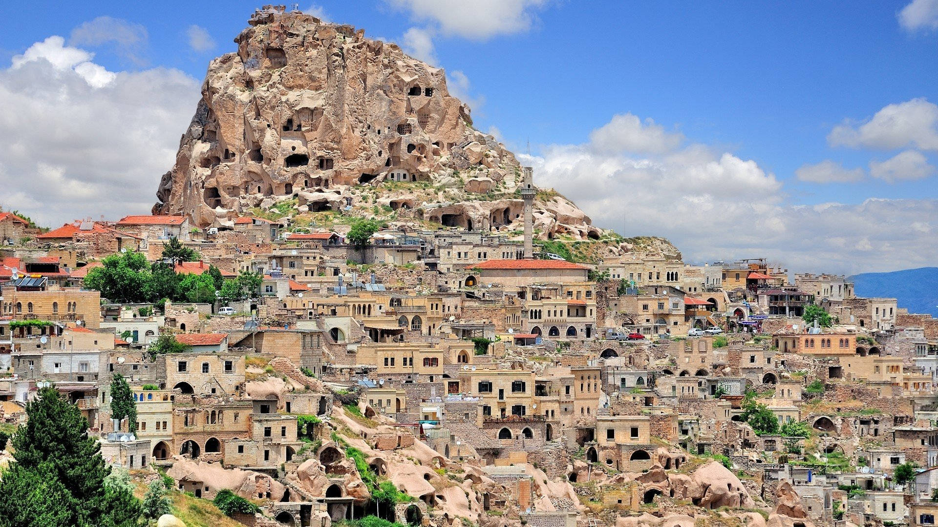 Cappadocia 桌布 壁紙 — 一個陽光普照的山丘小鎮，擁有赤陶屋頂的建築物，雕刻在巨大的岩石露頭上，藍天白雲。
