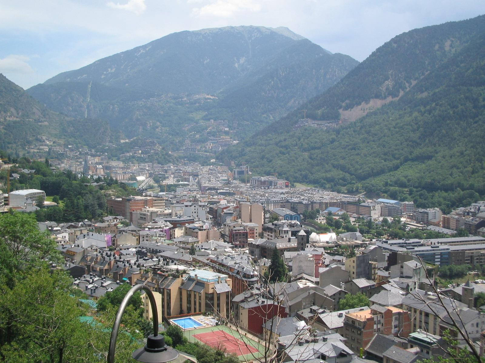山景 桌布 壁紙 — 一個陽光普照的山城，坐落在山谷中，四周是茂密的綠色森林和聳立的山峰，天空部分多雲。