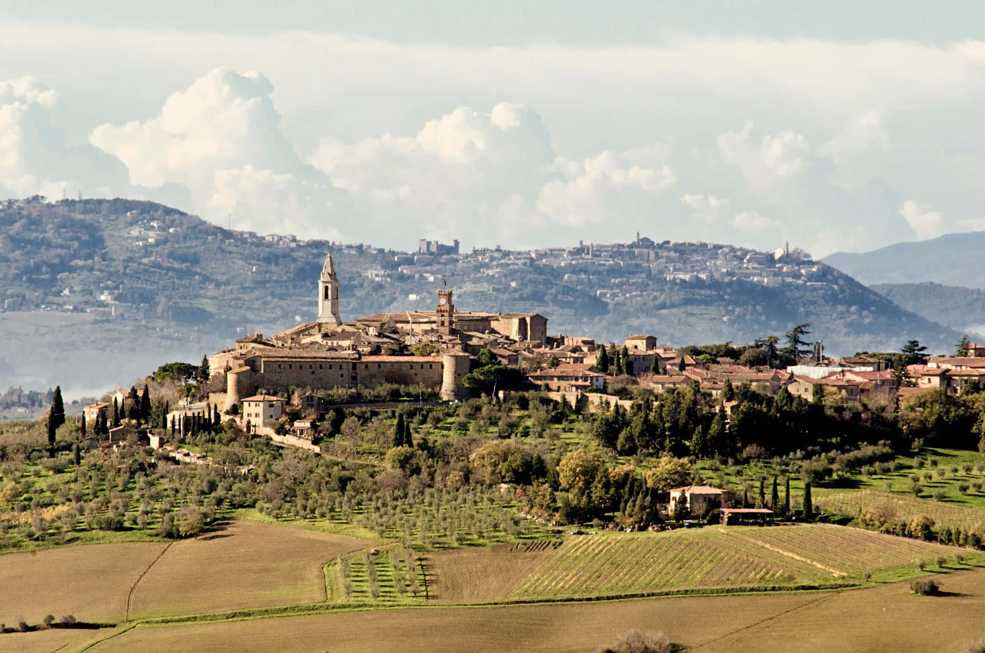 Tuscany 桌布 壁紙 — 一個陽光普照的山頂村莊，擁有陶土屋頂和石塔，俯瞰著多雲天空下連綿起伏的綠色田野。