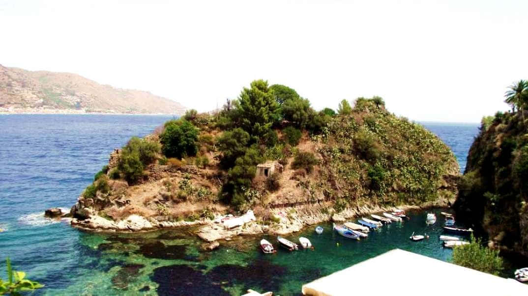 Italian Island 桌布 壁紙 — 一個陽光普照的岩石島嶼，綠意盎然，小船停泊在清澈見底的碧綠海水中。