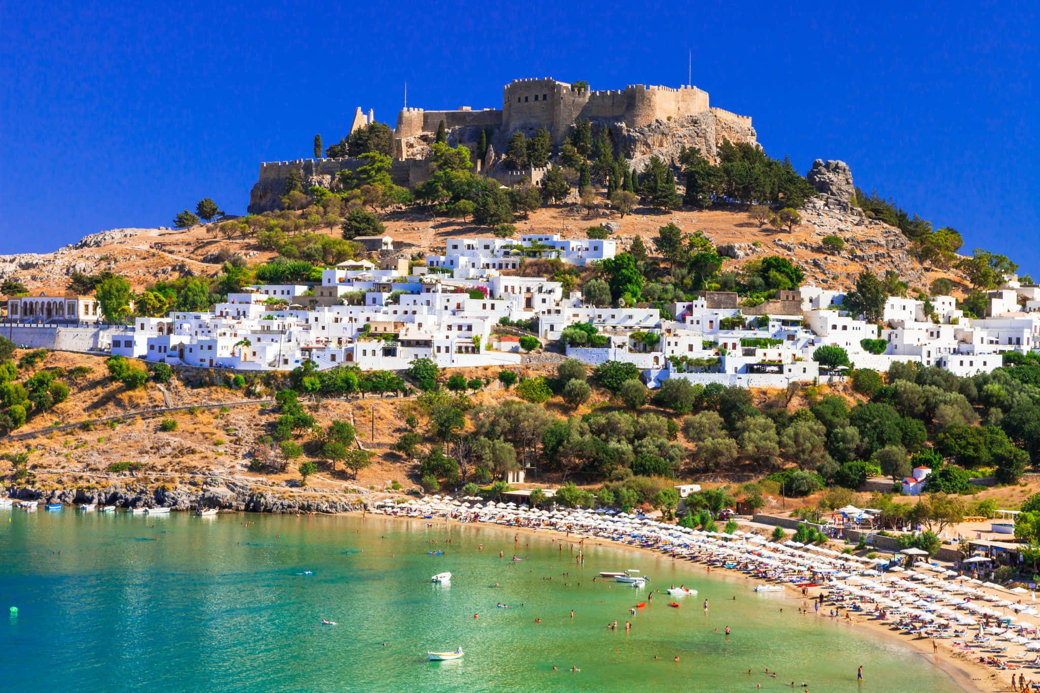 Mediterranean Beach 桌布 壁紙 — 一個陽光普照的希臘沿海村莊，白色建築沿著山坡層疊而下，通往點綴著船隻和日光浴者的碧綠海灣。