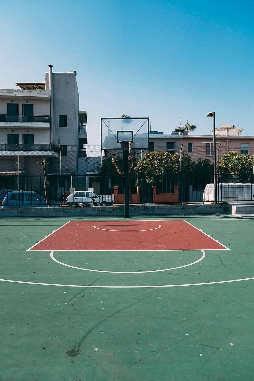 Basketball Court 桌布 壁紙 — 一個陽光普照的戶外籃球場，紅色和綠色的塗漆表面和白色線條，在晴朗的藍天下。
