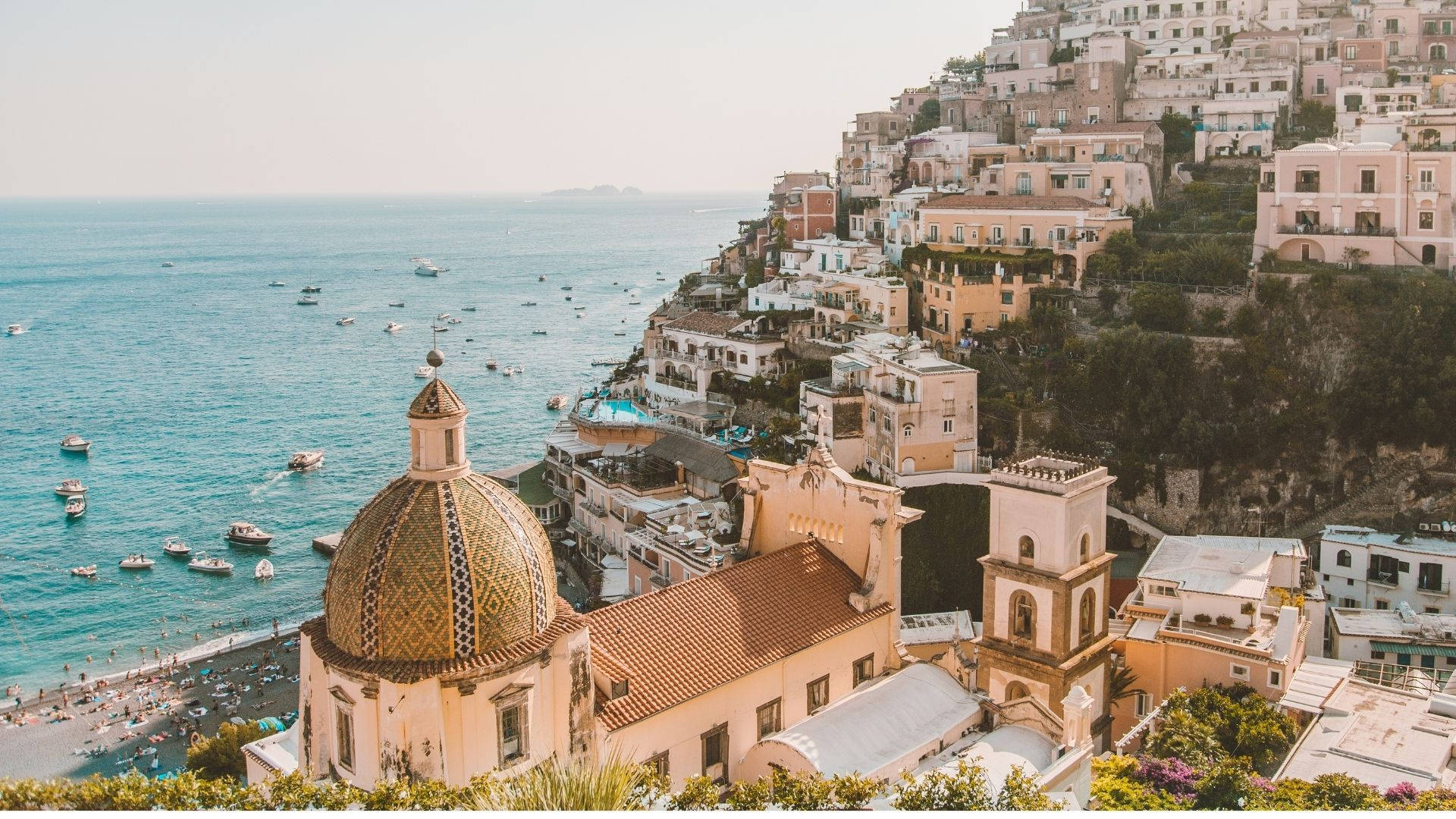 Amalfi Coast 桌布 壁紙 — 一個陽光普照的海岸小鎮緊貼在懸崖邊，俯瞰著點綴著船隻的碧綠海水和下方的沙灘。