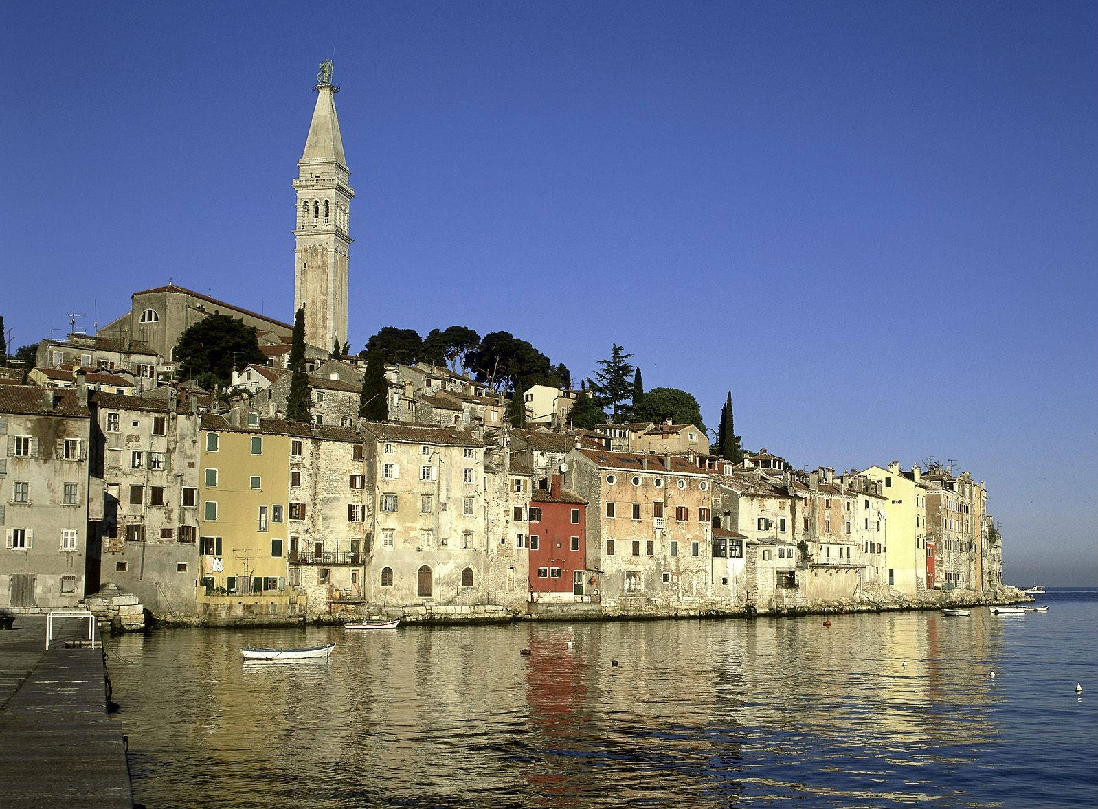 Croatia 桌布 壁紙 — 一個陽光普照的海濱小鎮，擁有粉彩建築和高聳的鐘樓，倒映在平靜的藍色水面上。