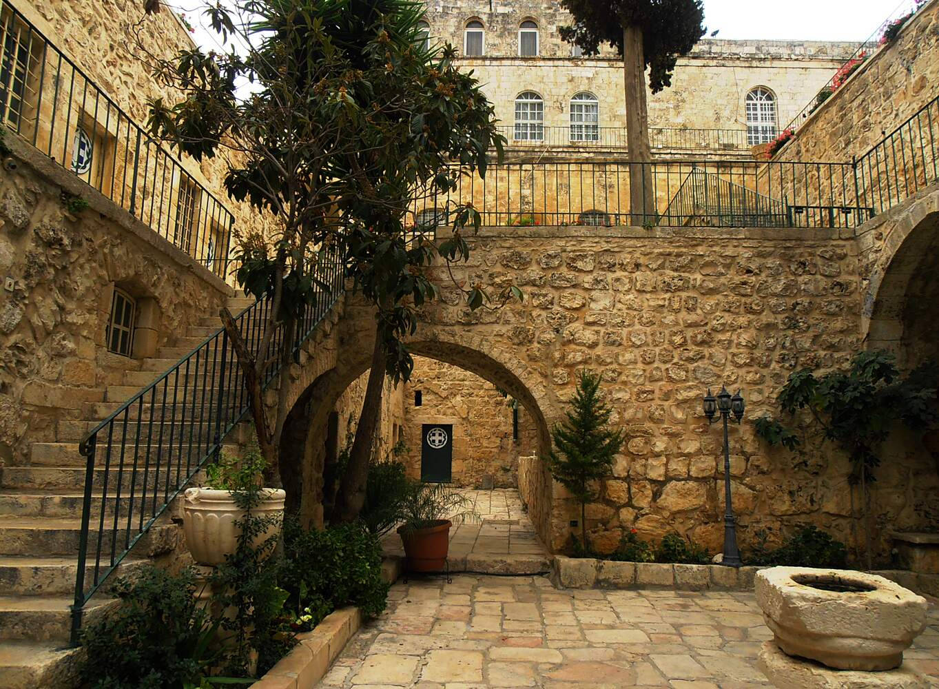 Israel 桌布 壁紙 — 一個陽光普照的石頭庭院，拱形通道、樓梯和盆栽樹木點綴在古老建築之間。