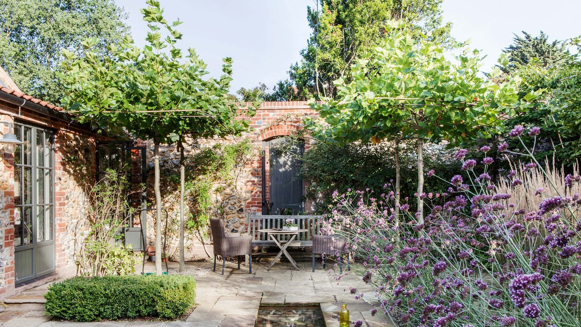 Cottage Garden 桌布 壁紙 — 一個陽光普照的石頭庭院花園，種有薰衣草灌木、木桌椅子，以及攀爬在磚牆上的綠色植物。