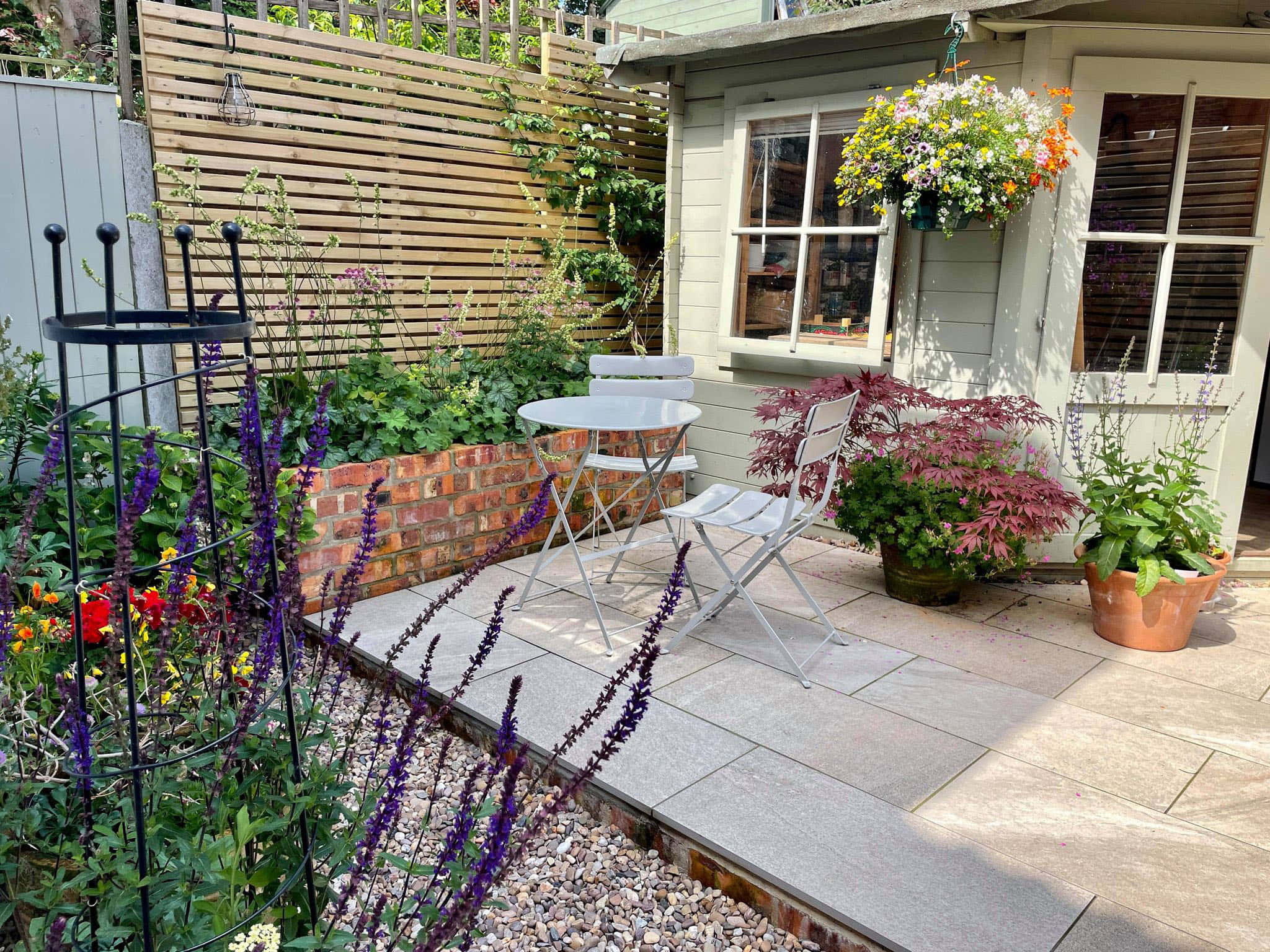 Cottage Garden 桌布 壁紙 — 一個陽光普照的花園露台，設有白色小酒館套裝，周圍環繞著充滿活力的紫色花朵、盆栽植物和木柵欄。