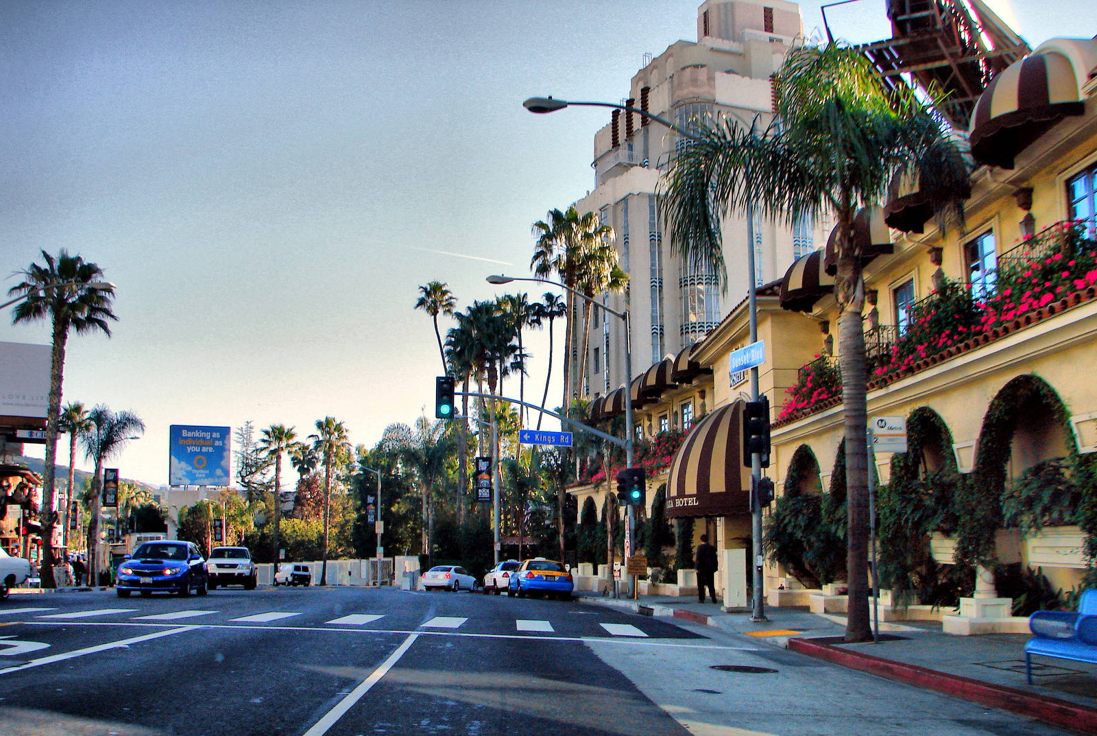 hollywood street 桌布 壁紙 — 一個陽光普照的街角，位於棕櫚樹成蔭的市區，有一座宏偉的酒店和汽車在交通燈前等候。