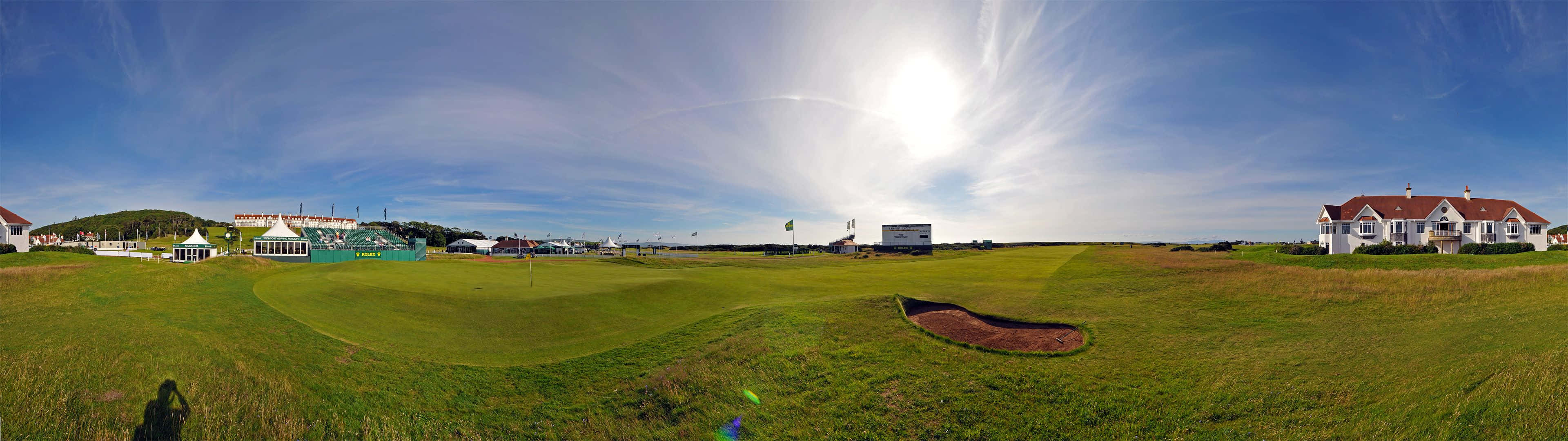 Golf 3840x1080 桌布 壁紙 — 一個陽光普照的高爾夫球場全景，擁有充滿活力的綠色球道、沙坑和遠處的建築物，在清澈的藍天下。
