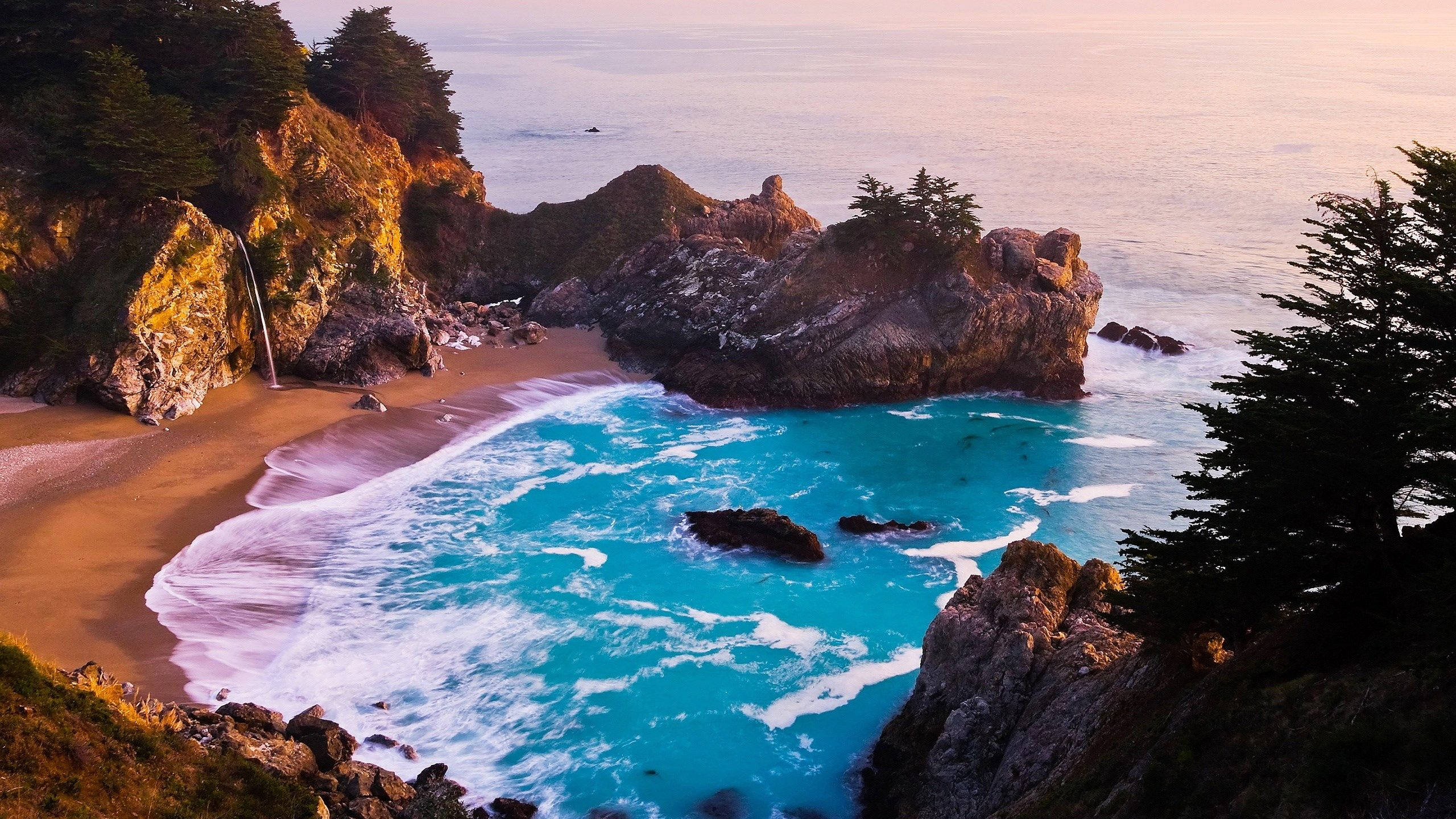 Malibu California 桌布 壁紙 — 一個隱蔽的小海灣，在日落時分，碧綠的海浪拍打著金色的沙灘，四周環繞著崎嶇的懸崖和常綠樹木。