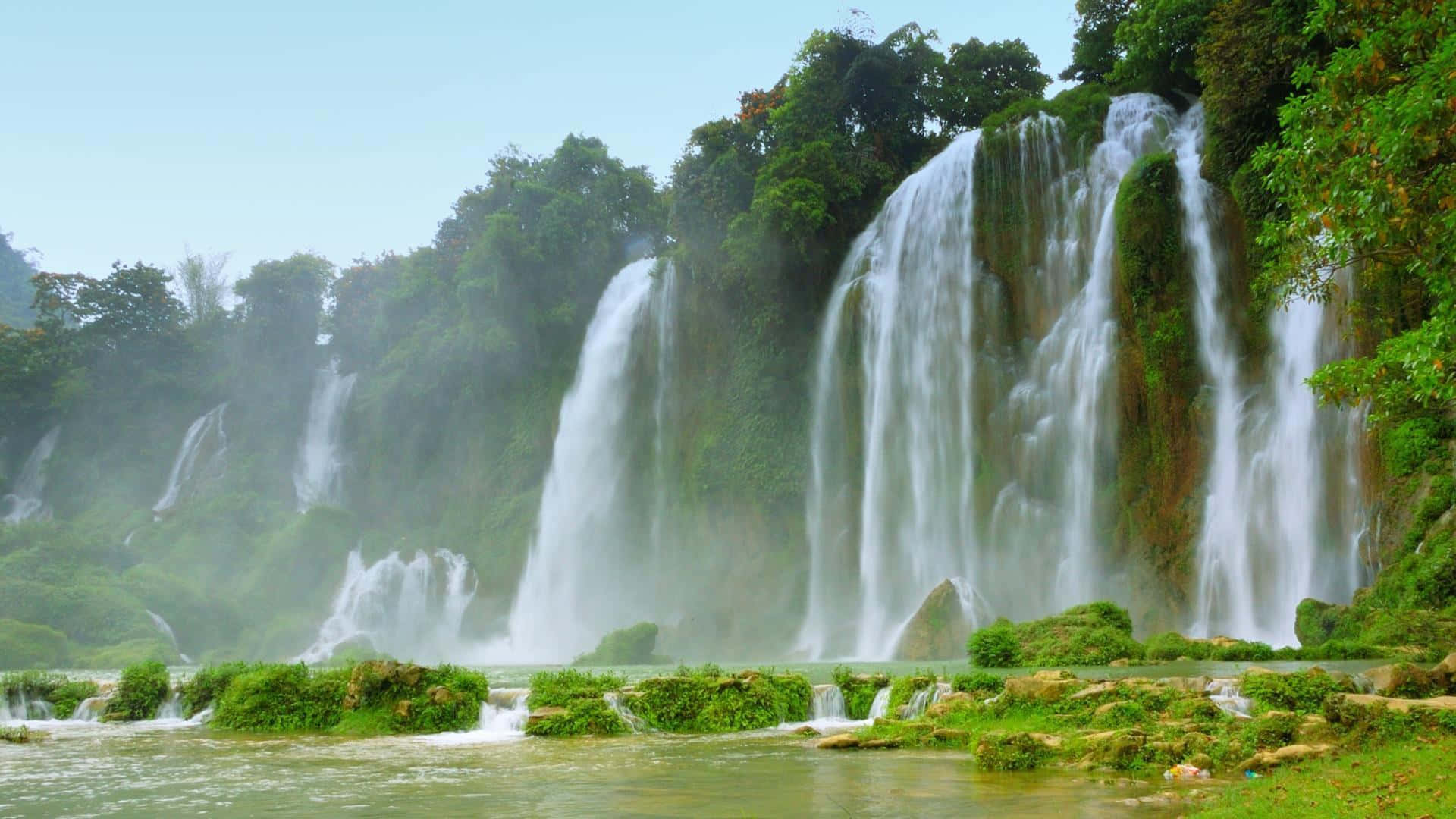 3d Waterfall 桌布 壁紙 — 一個雄偉的多層瀑布穿過茂密的綠色叢林葉片，傾瀉而下，流入下方霧氣瀰漫的水潭。