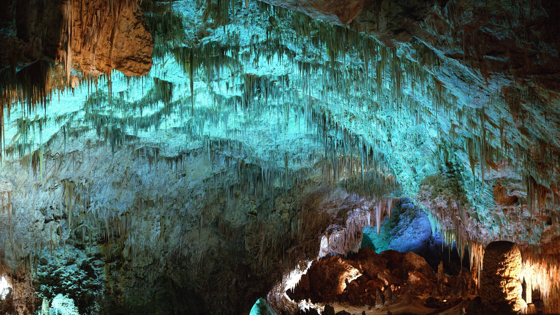 Carlsbad Caverns 桌布 壁紙 — 一個雄偉的洞穴，以綠松石和琥珀色燈光照明，突顯出複雜的鐘乳石和石筍。
