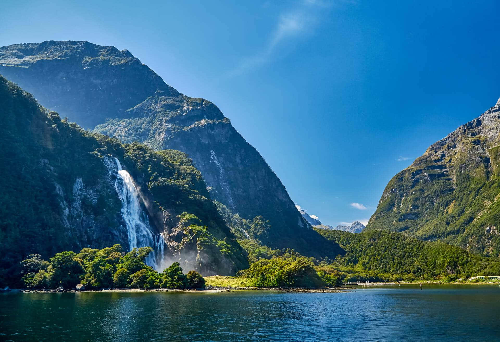 Beautiful Mountain Lake 桌布 壁紙 — 一個雄偉的瀑布從鬱鬱蔥蔥的綠色山脈傾瀉而下，流入晴朗天空下平靜的藍色峽灣。