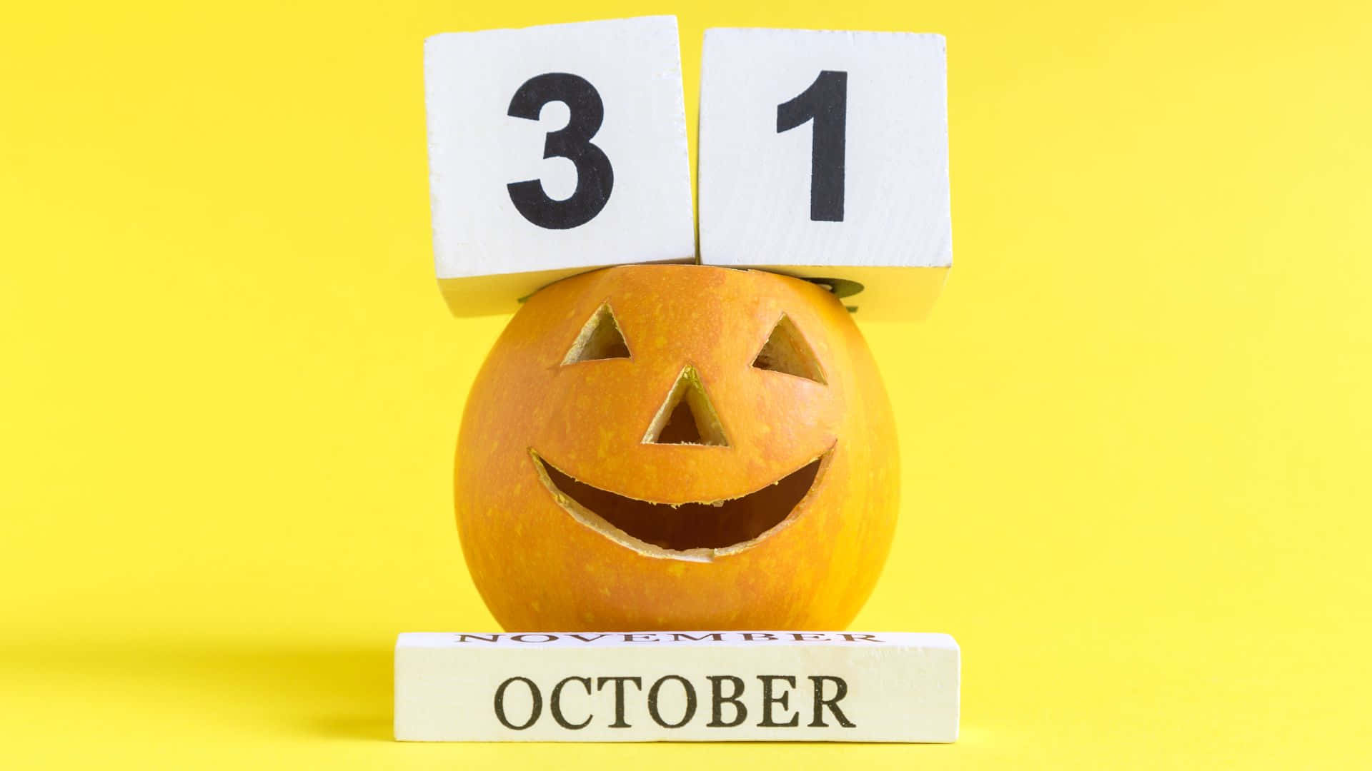 October 31st 桌布 壁紙 — 一個雕刻成笑臉的橙色南瓜，平衡地疊著木塊，上面顯示日期31，下面顯示October，背景是鮮黃色。