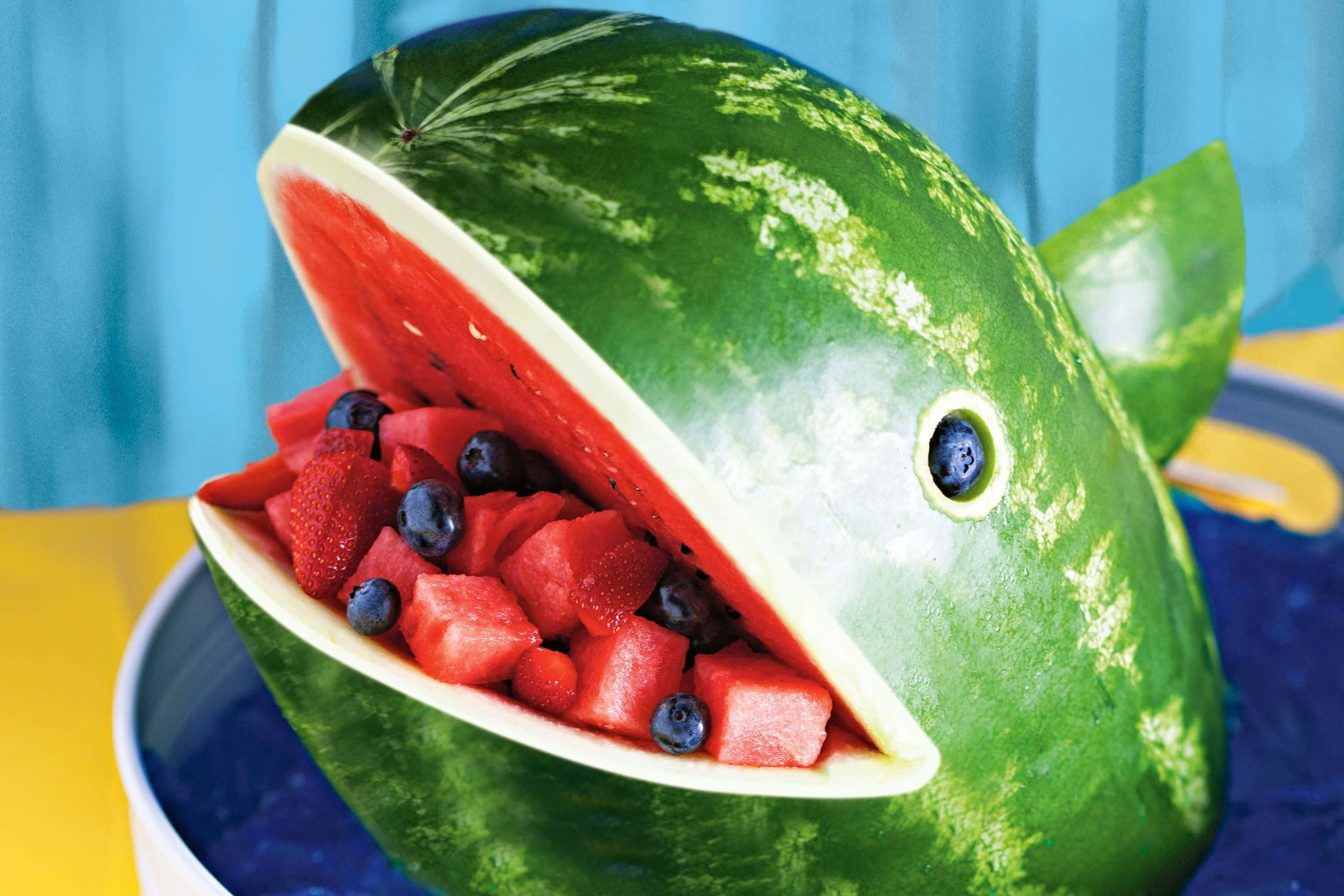 Watermelon 桌布 壁紙 — 一個雕刻成魚形、張開嘴巴的西瓜，裡面裝滿了西瓜丁、草莓和藍莓。