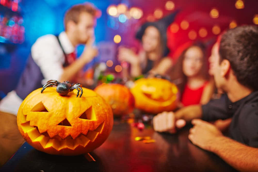 Halloween Pumpkin 桌布 壁紙 — 一個雕刻的傑克南瓜燈上有一隻塑膠蜘蛛，放在前景，朋友們在昏暗的派對環境中慶祝萬聖節。