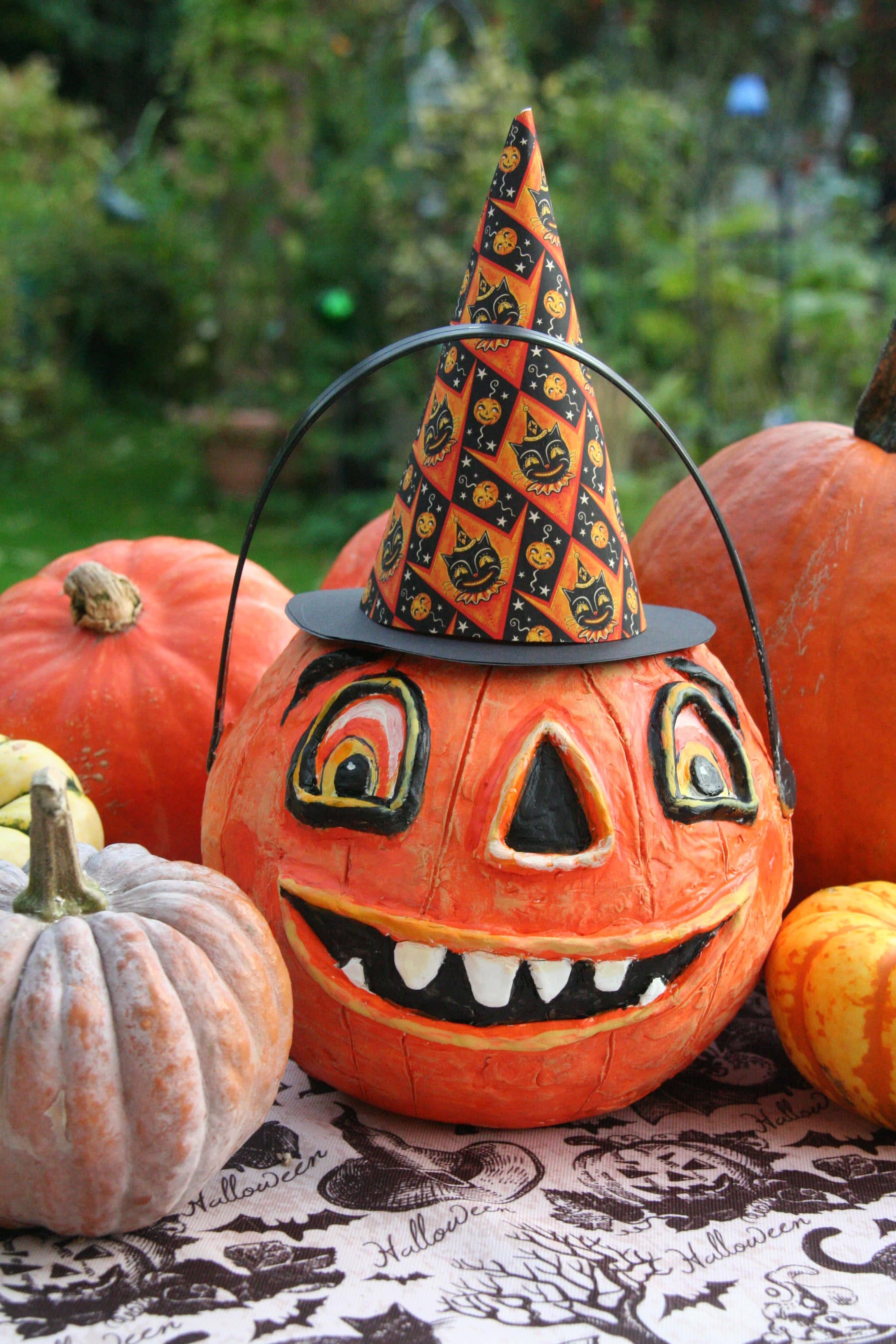 jack o'lantern 桌布 壁紙 — 一個雕刻的傑克南瓜燈戴著有圖案的女巫帽，坐在萬聖節主題桌布上的南瓜之間。