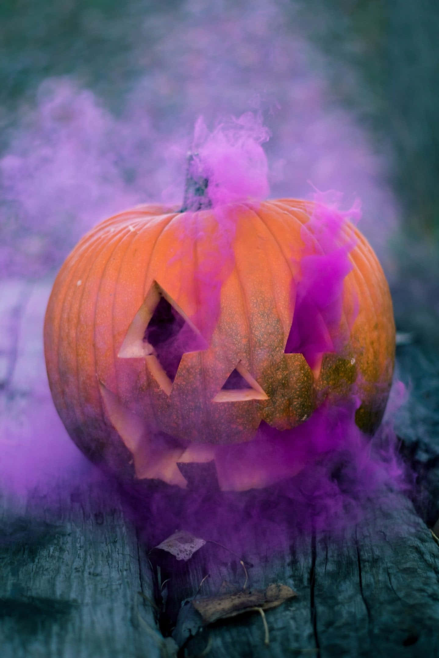 Fall Halloween iPhone 桌布 壁紙 — 一個雕刻的橙色南瓜，帶有三角形眼睛和鋸齒狀的笑容，在深藍綠色背景下散發出充滿活力的紫色煙霧。