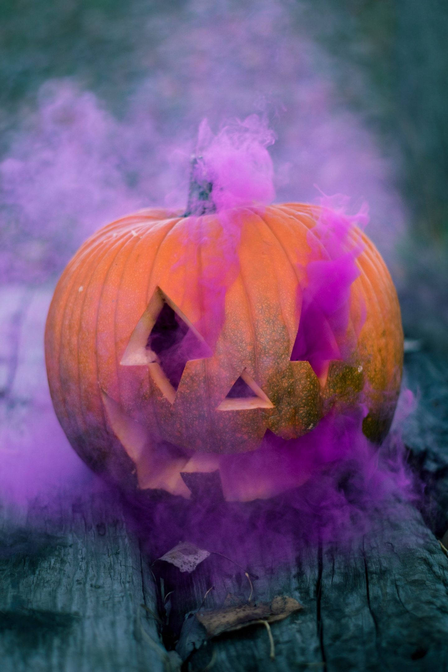Cute Halloween Phone 桌布 壁紙 — 一個雕刻的橙色南瓜，帶有三角形眼睛和鋸齒狀嘴巴，在深色木質表面上散發出鮮豔的紫色煙霧。