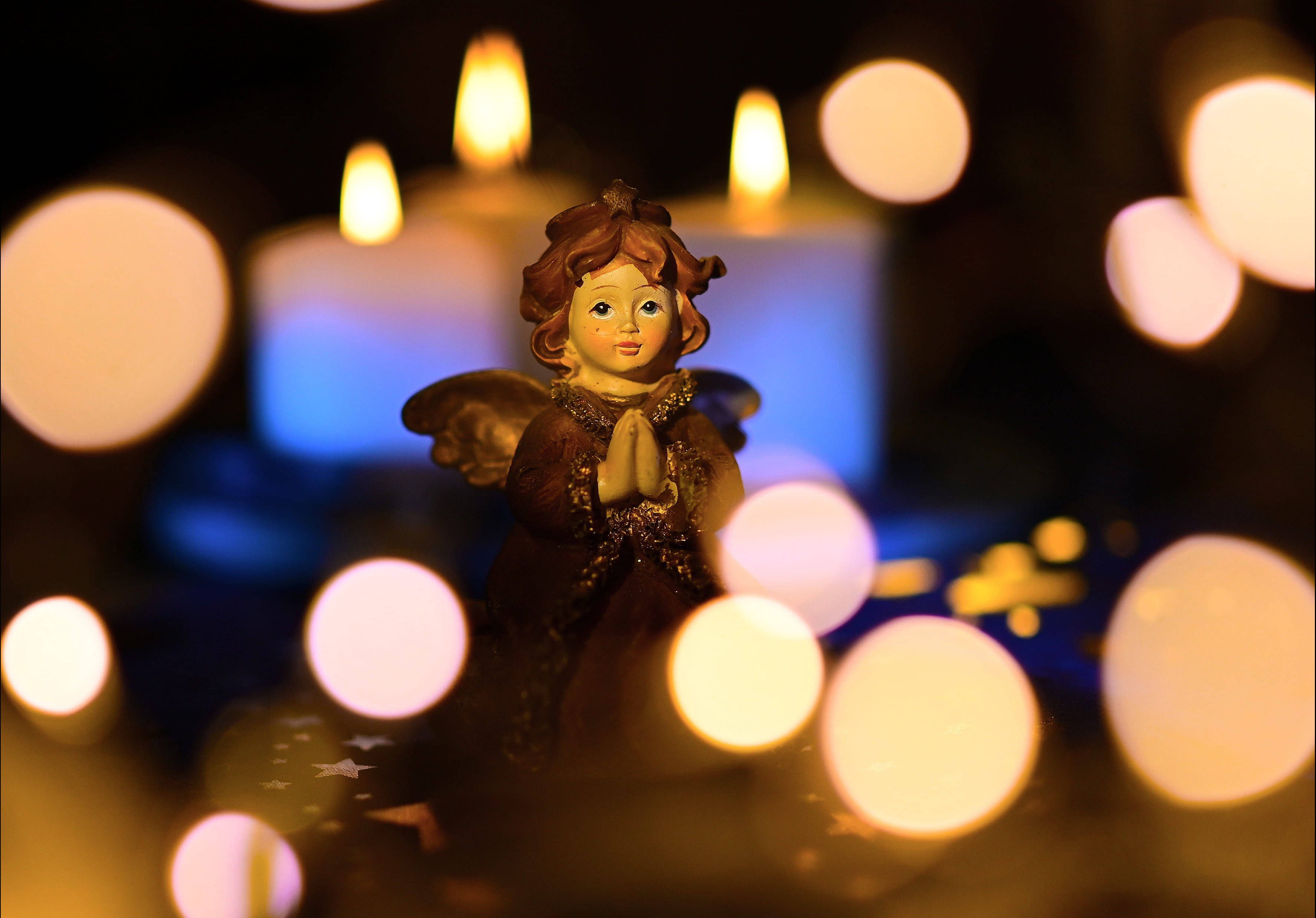 angels 桌布 壁紙 — 一個雙手合十的小天使雕像，坐在昏暗的環境中，周圍是閃爍的燭光和柔和的散景。