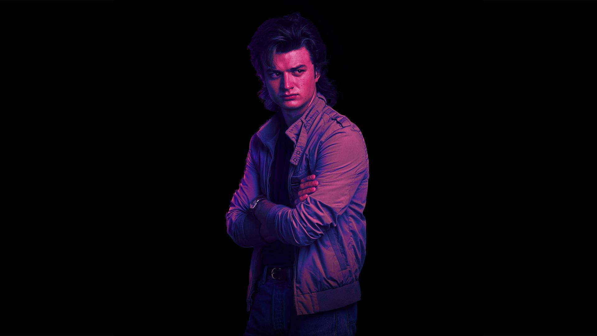 Joe Keery 桌布 壁紙 — 一個雙臂交叉的年輕男子站在黑色背景前，沐浴在戲劇性的紫色和粉紅色霓虹燈光中。