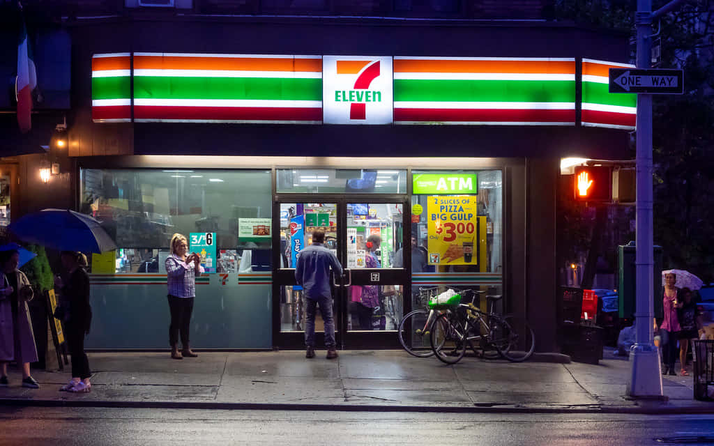 7-ELEVEN 桌布 壁紙 — 一個雨夜場景，在一間燈火通明的7-Eleven商店外，有人撐著雨傘，附近停著一輛自行車。