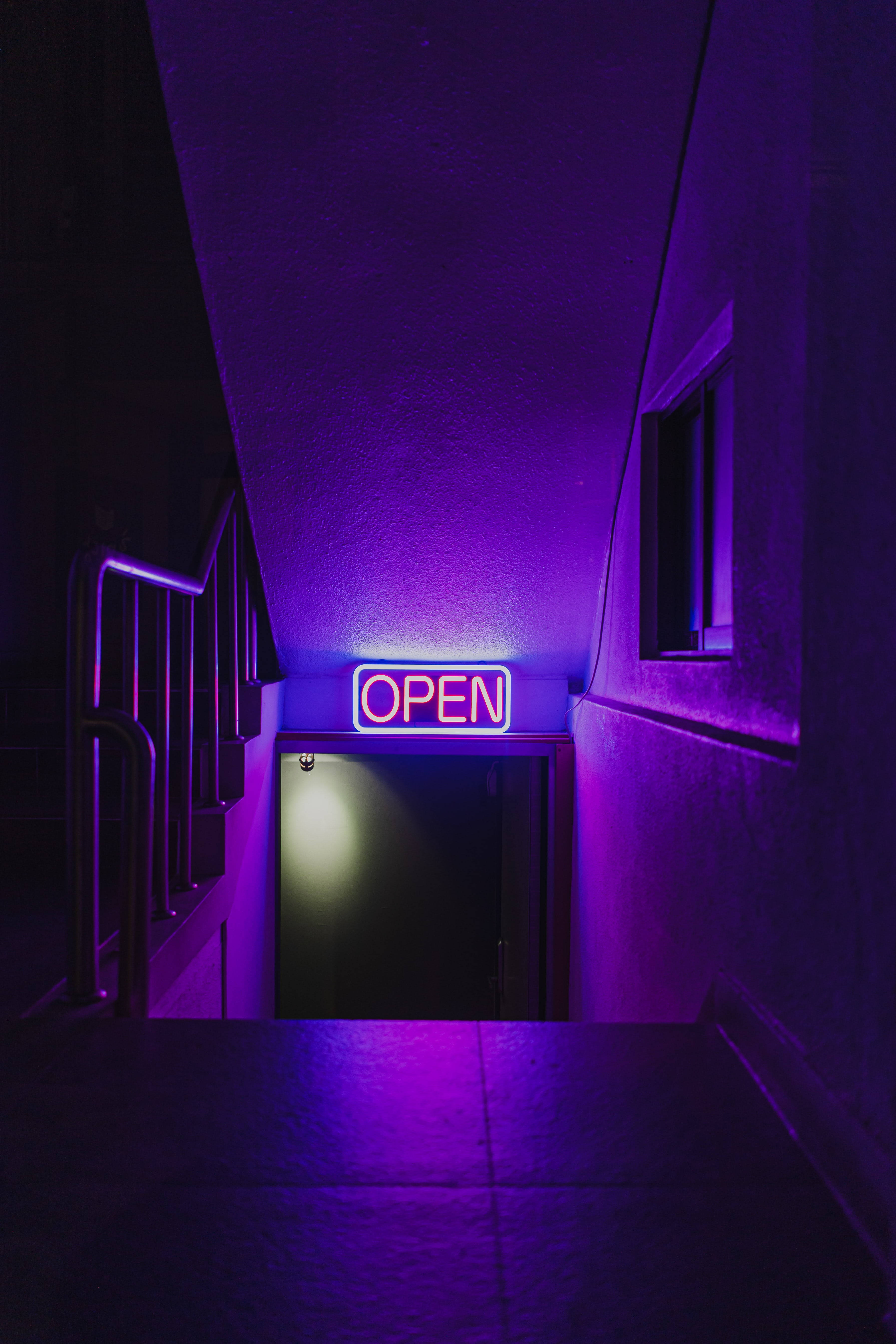 Dark Neon 桌布 壁紙 — 一個霓虹燈「營業中」標誌在昏暗的樓梯間發出紫色光芒，將鮮豔的光線投射在混凝土牆壁和金屬欄杆上。