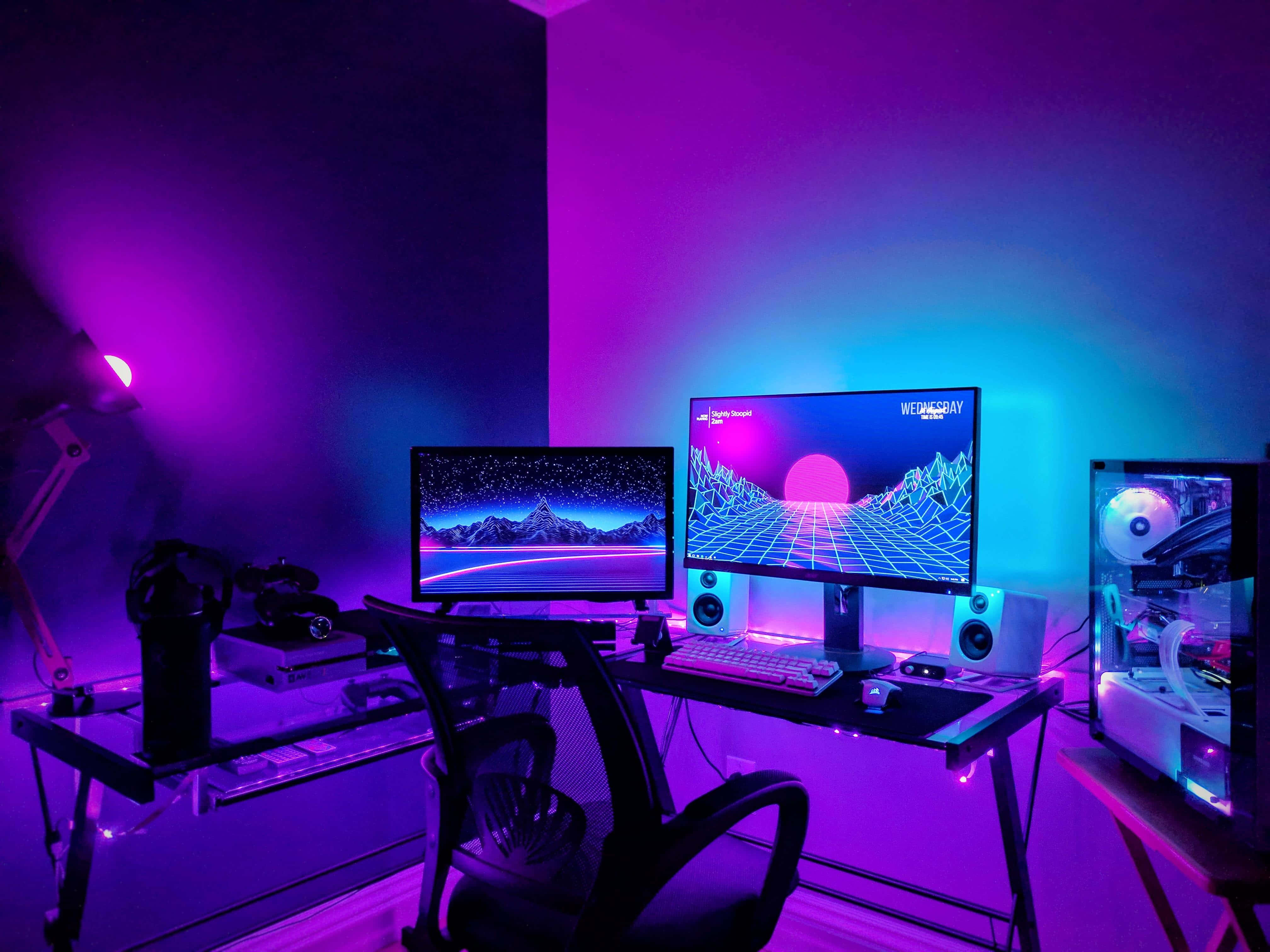 Gaming Pc Setup 桌布 壁紙 — 一個霓虹燈遊戲設置，配備雙顯示器，在紫色和藍色環境光下顯示復古未來主義景觀。