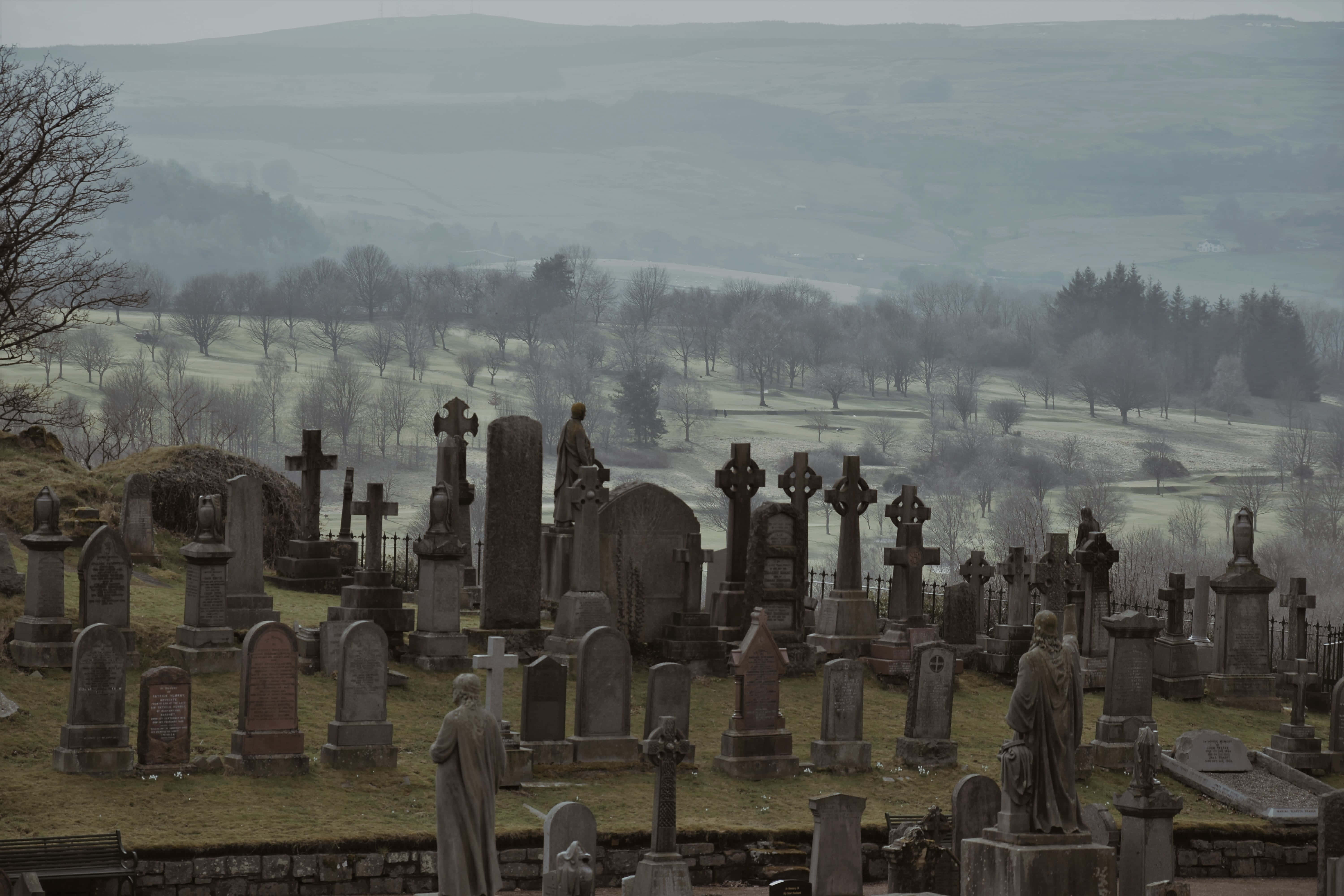 Gravestone 桌布 壁紙 — 一個霧氣瀰漫的墓地，佈滿風化的石十字架和雕像，俯瞰著灰色天空下連綿起伏的綠色山谷。