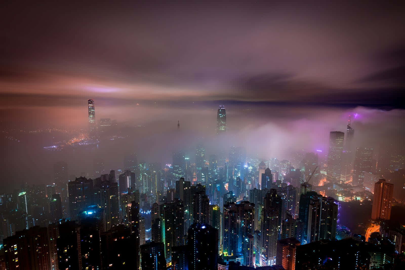 City At Night 桌布 壁紙 — 一個霧氣瀰漫的夜晚城市景觀，摩天大樓在紫色和藍色霓虹燈光下閃耀，天空多雲。