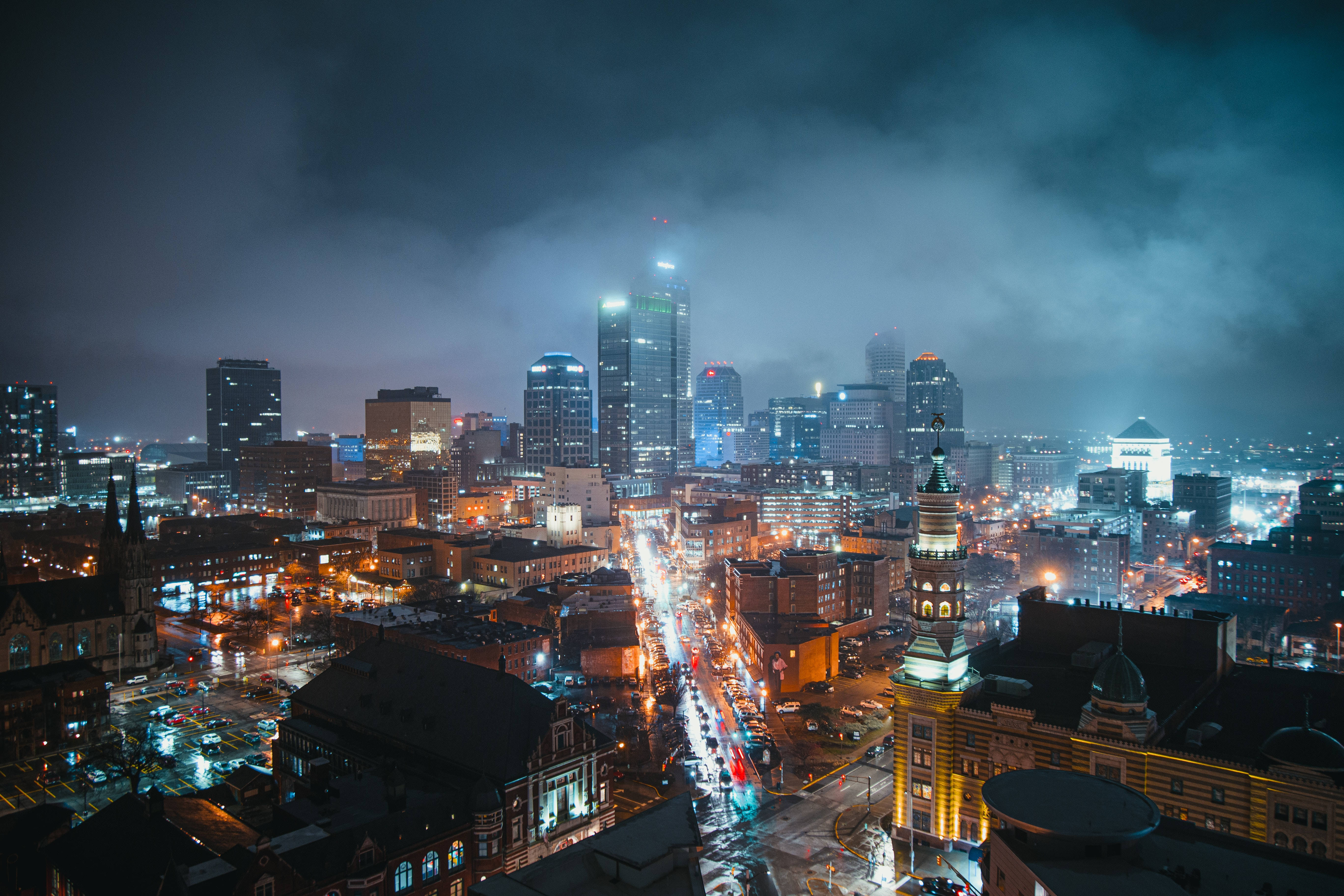雲朵 桌布 壁紙 — 一個霧氣瀰漫的夜晚城市景觀，閃爍的摩天大樓和濕漉漉的街道在陰雲密布的天空下反射出鮮豔的路燈。