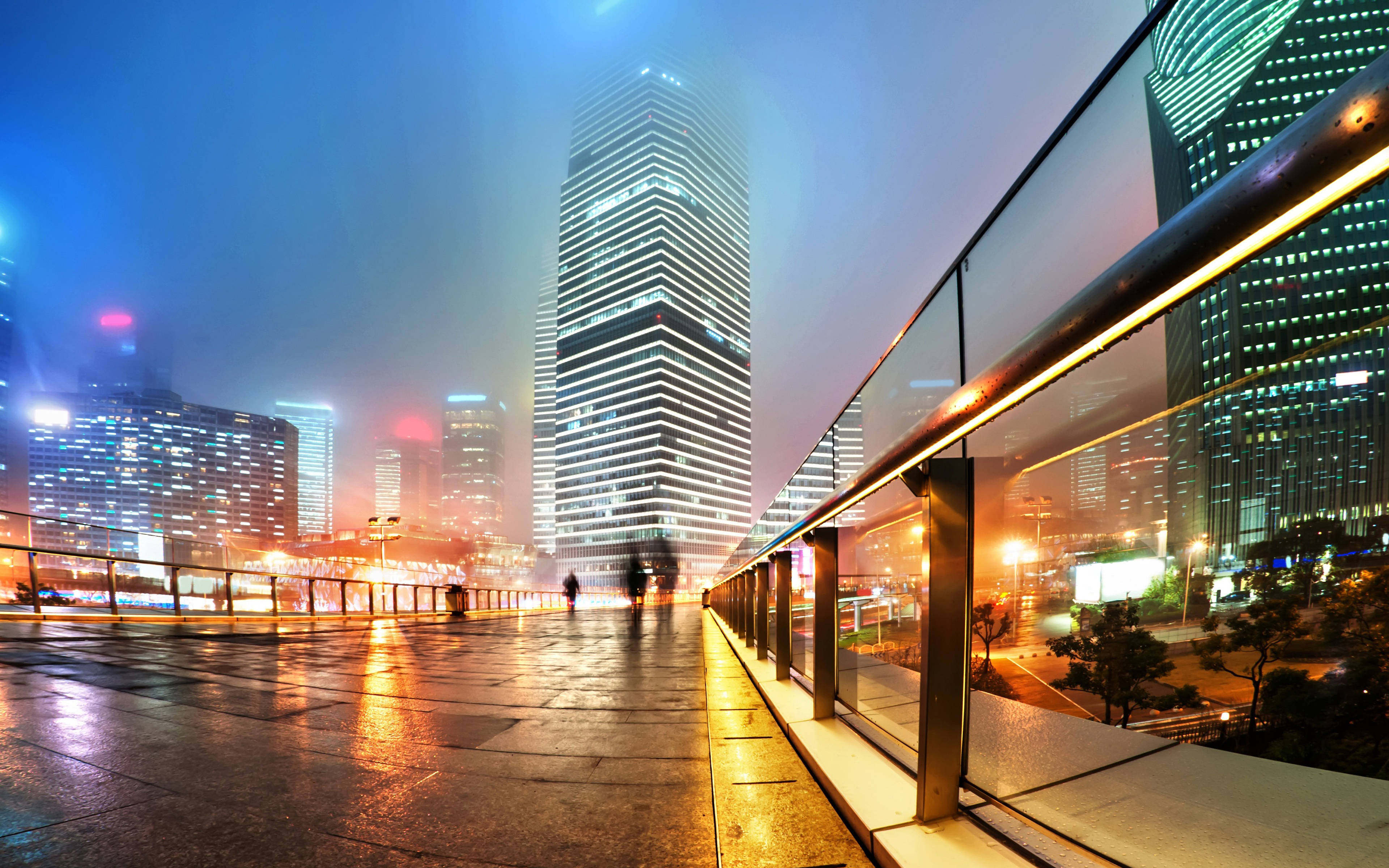 Shanghai 桌布 壁紙 — 一個霧氣瀰漫的夜晚城市景觀，高聳的摩天大樓在霓虹燈下閃耀，燈光反射在潮濕的路面上。