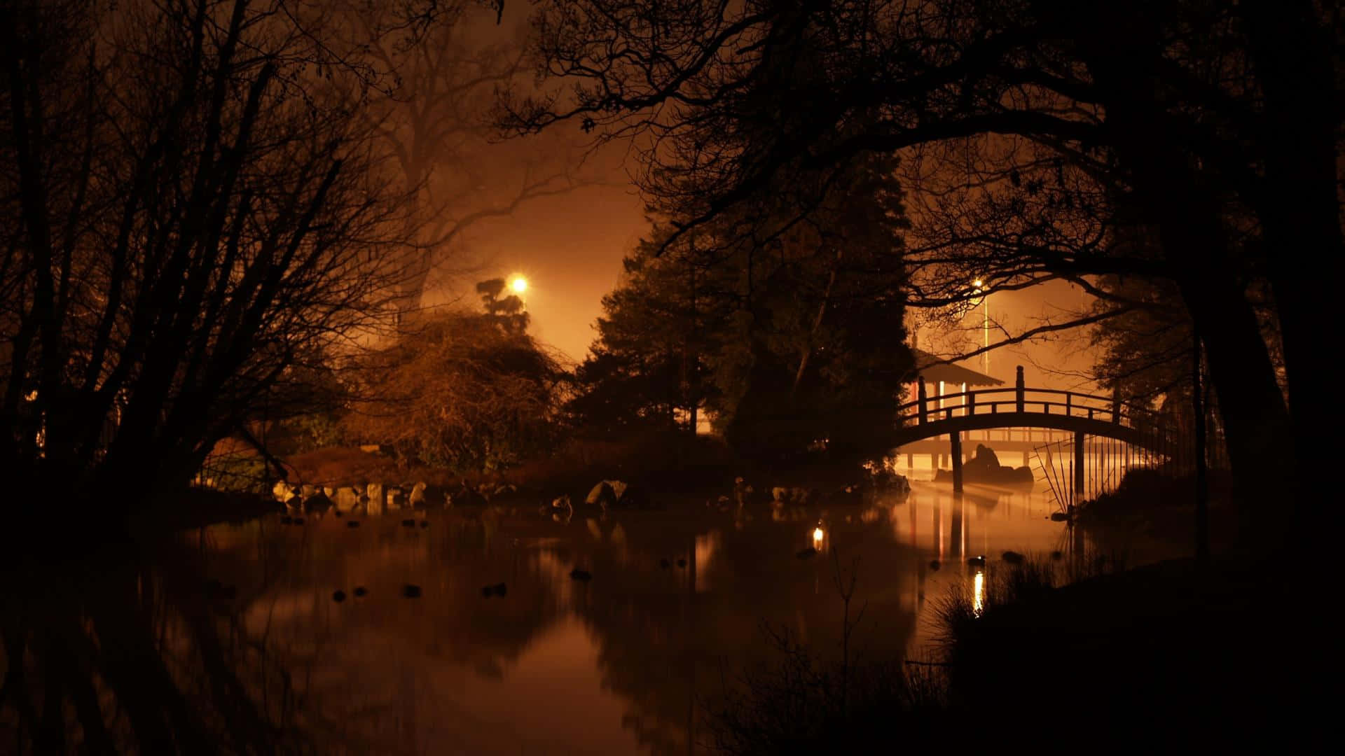 日式暗黑 桌布 壁紙 — 一個霧氣瀰漫的夜晚場景，木橋橫跨平靜的池塘，倒映著樹木和街燈溫暖的橙色燈光。