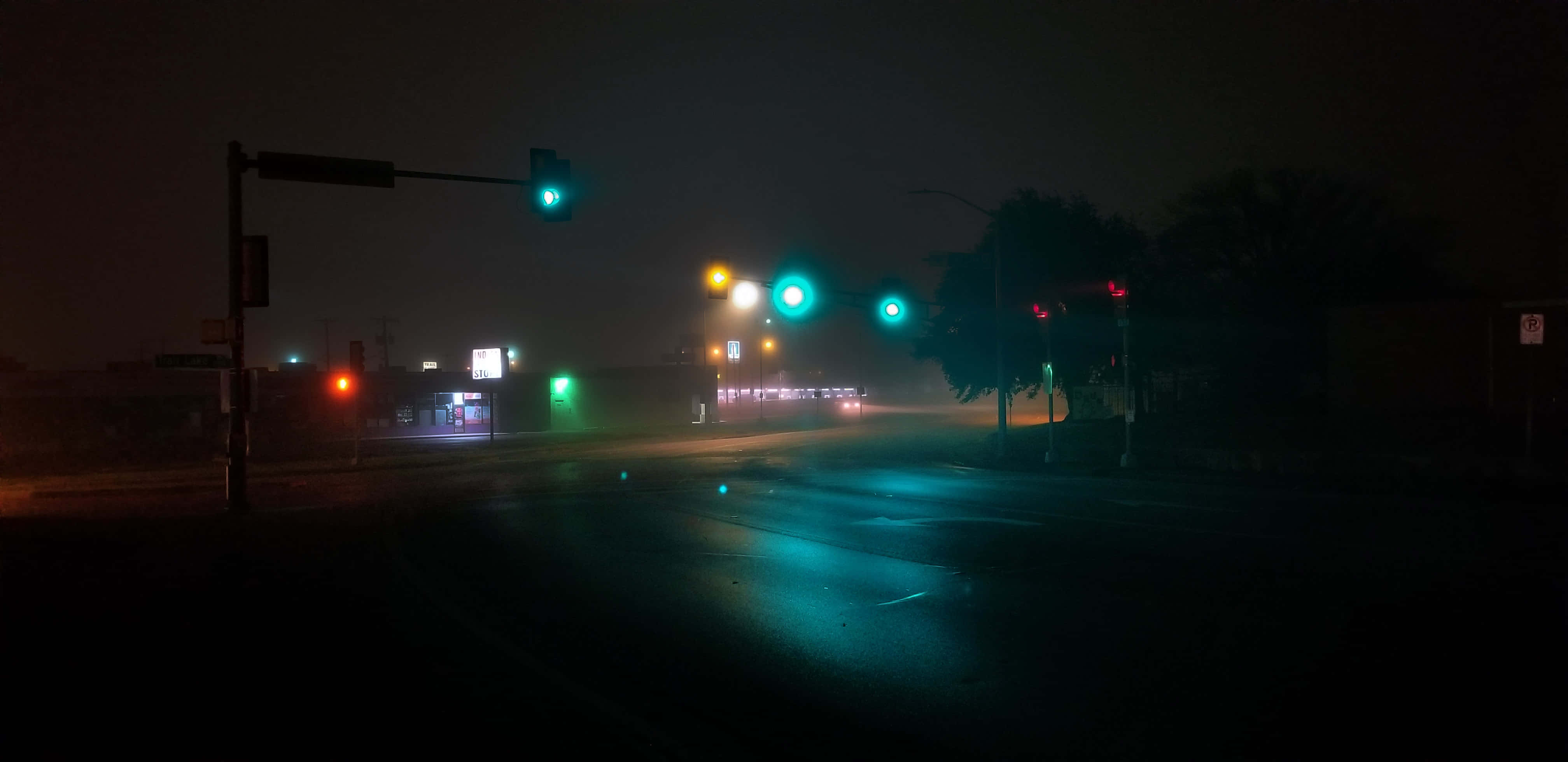 Doomer 桌布 壁紙 — 一個霧氣瀰漫的夜晚街道十字路口，被發光的交通號誌燈照亮，在濕漉漉的路面上投射出青色倒影。