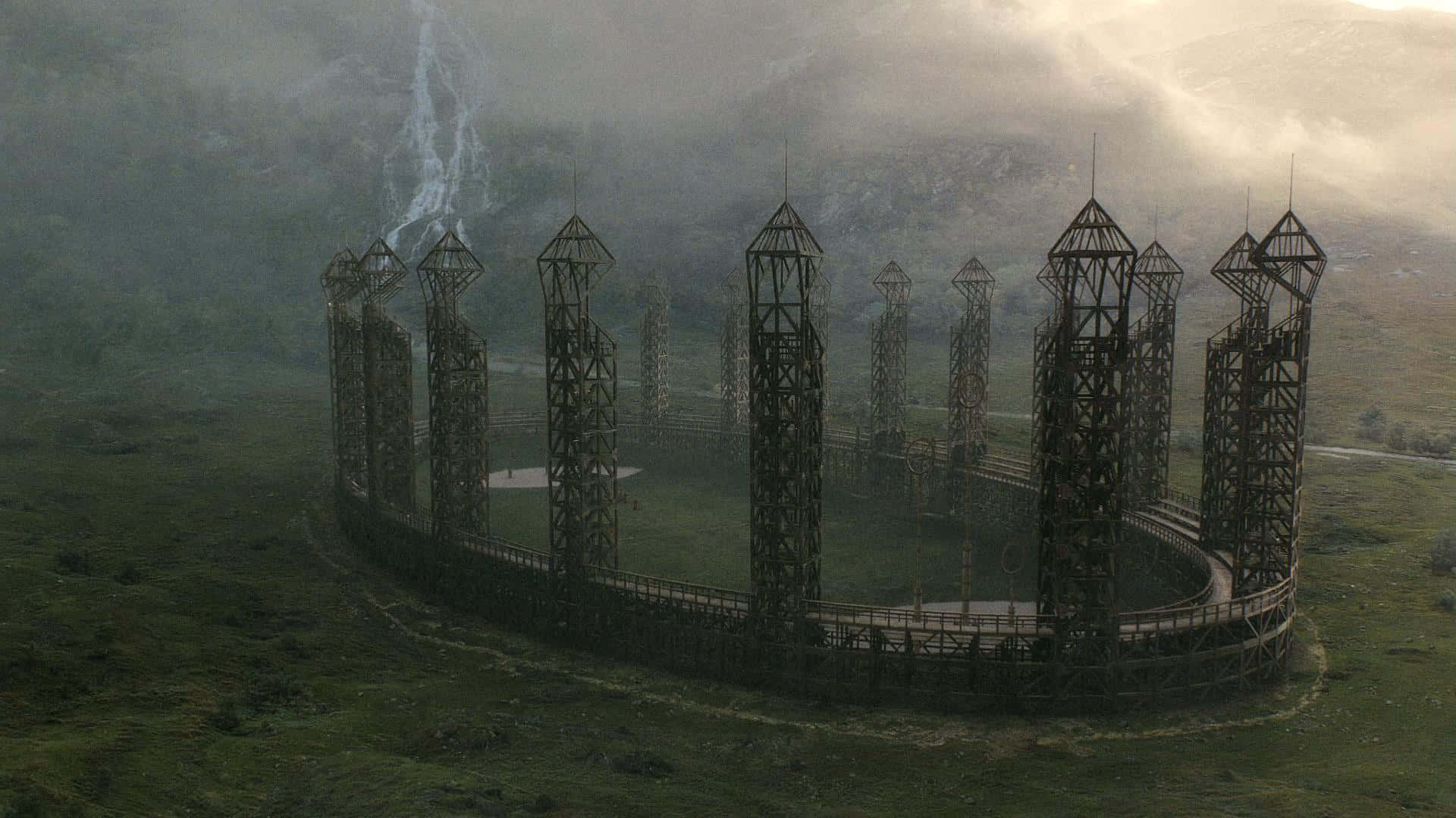 Quidditch 桌布 壁紙 — 一個霧氣瀰漫的山景，有一個巨大的圓形木結構，帶有高高的尖頂和一個中央平台。