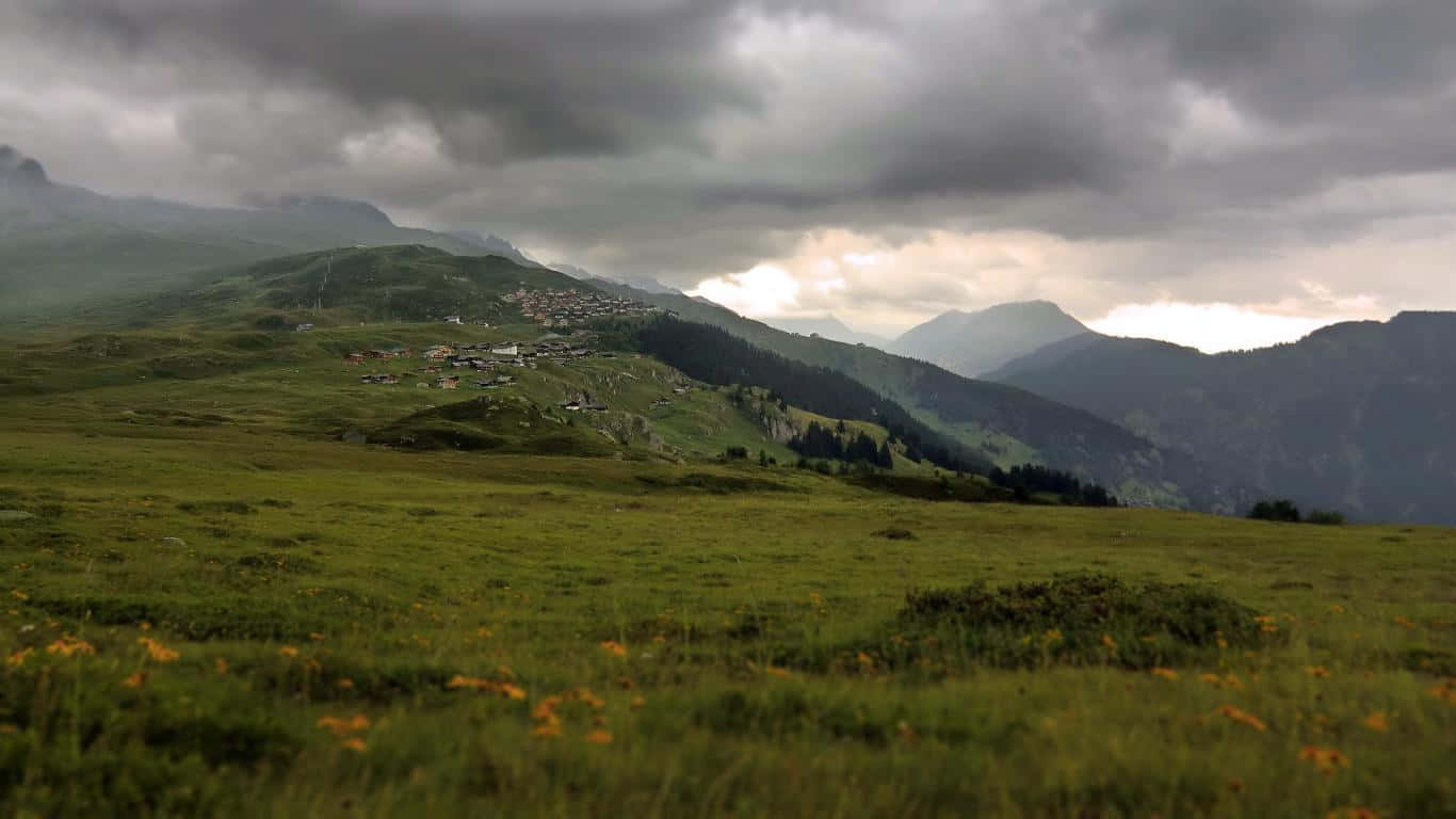 Weather 桌布 壁紙 — 一個霧氣瀰漫的山谷，一個小村莊坐落在綠色的山坡之間，上方是戲劇性的多雲天空。