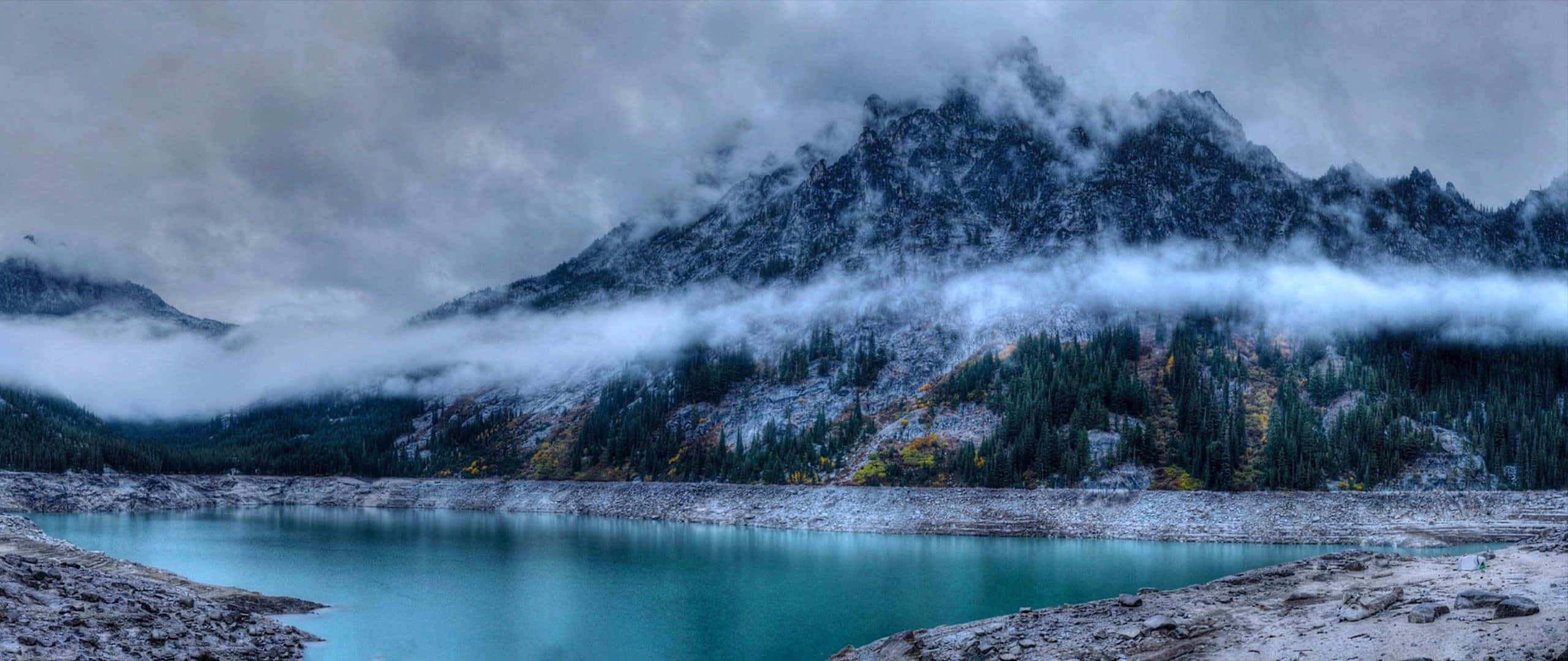 山景 桌布 壁紙 — 一個霧氣瀰漫的山間湖泊，在積雪的山峰和 swirling 的雲層下映照出碧綠色的湖水。