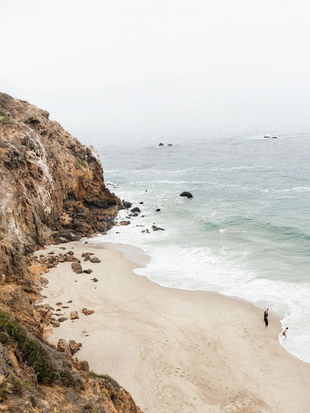 malibu iPhone 桌布 壁紙 — 一個霧氣瀰漫的海岸沙灘，有崎嶇的懸崖和溫柔的海浪拍打著岸邊，兩個人在水邊漫步。