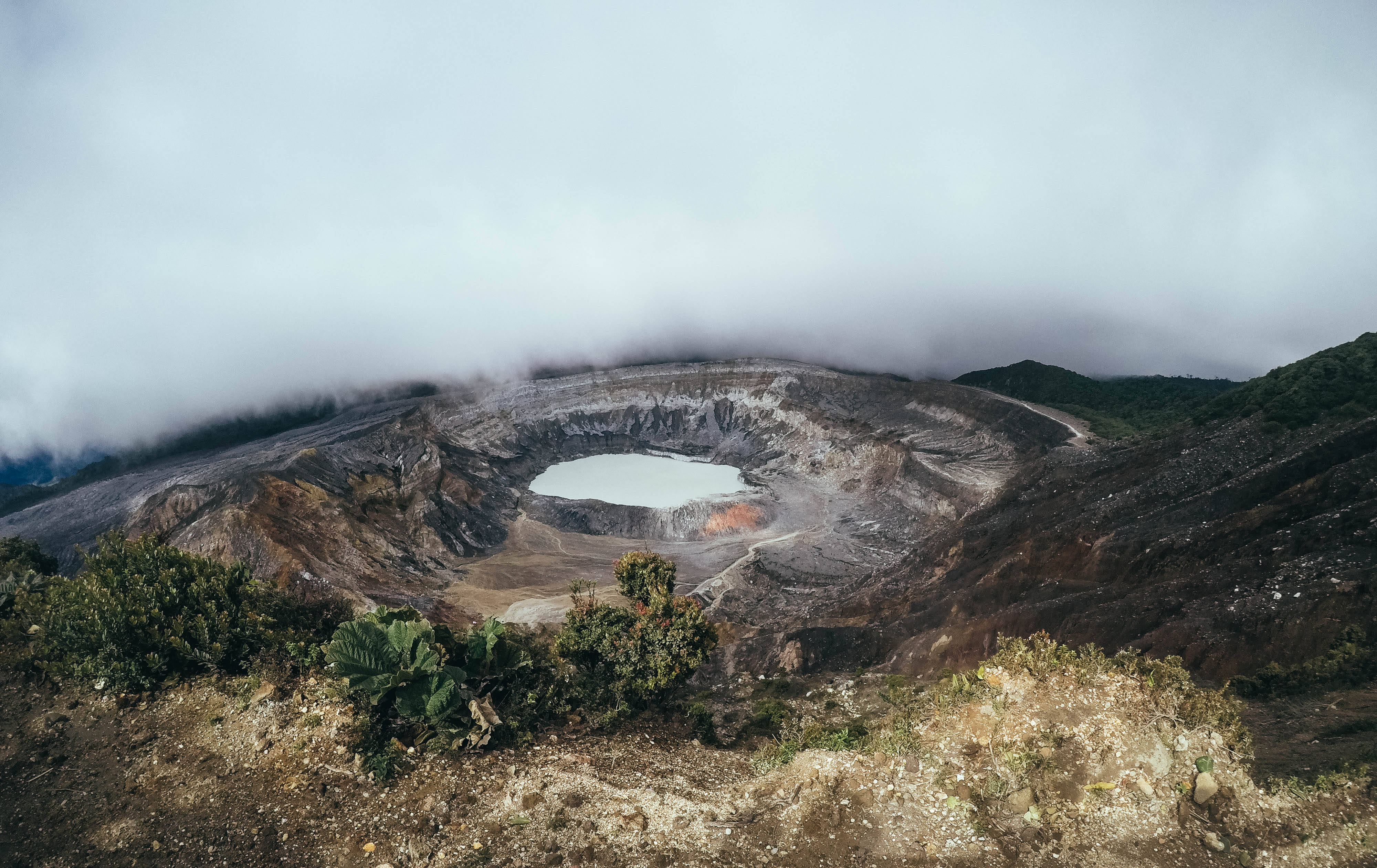 Costa 桌布 壁紙 — 一個霧氣瀰漫的火山口湖，坐落在崎嶇的地形中，邊緣長滿茂密的綠色植物，天空多雲。